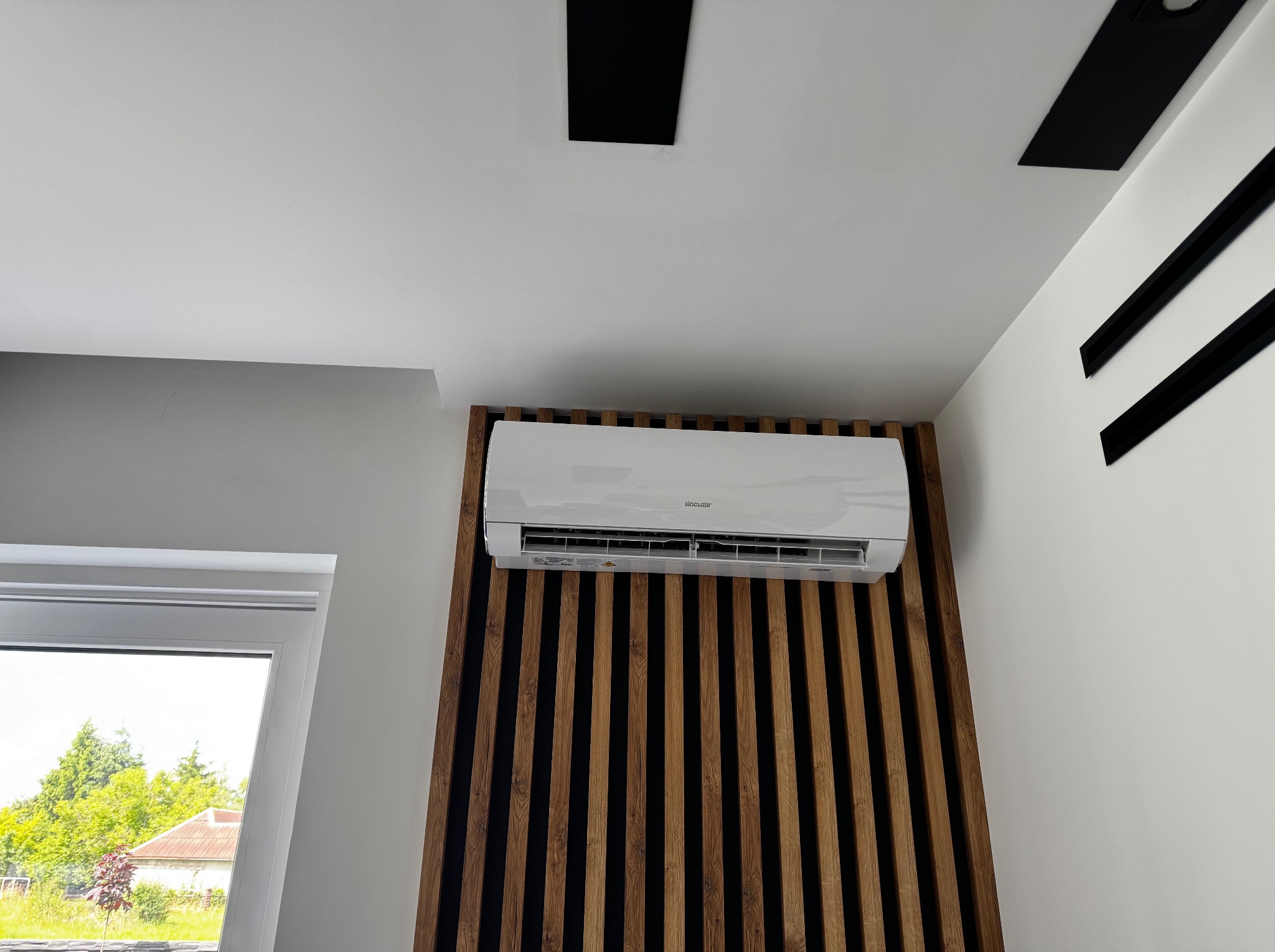 Biała klimatyzacja Sinclair na ścianie z drewnianymi lamelami. Widok z dołu na sufit z czarnymi elementami dekoracyjnymi. Okno z widokiem na zieleń.