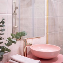 Paula Schleser - Różowa umywalka nablatowa w łazience z płytkami w odcieniach różu, złota bateria i detale, szklana kabina prysznicowa w tle oraz eukaliptus w wazonie.