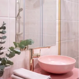 Różowa umywalka nablatowa w łazience z płytkami w odcieniach różu, złota bateria i detale, szklana kabina prysznicowa w tle oraz eukaliptus w wazonie.