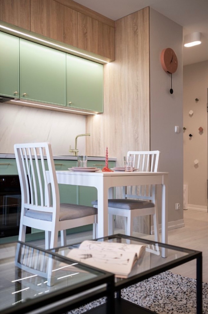 Jasna kuchnia z elementami drewna i pistacjowej zieleni, biały stół z krzesłami, na pierwszym planie szklany stolik kawowy z rozłożonym szkicem projektu