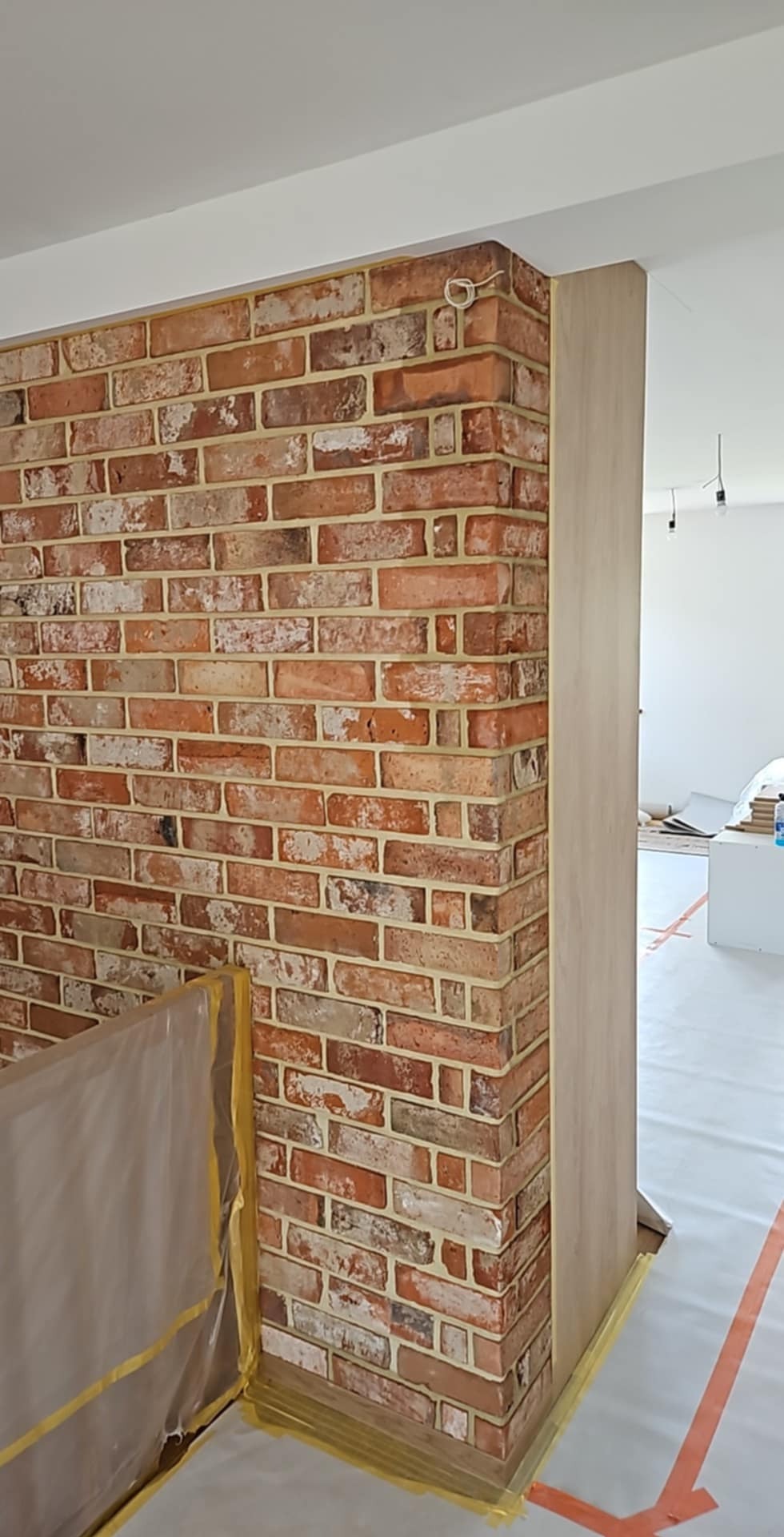 Wykończona ściana z cegły klinkierowej wewnątrz pomieszczenia, widoczna listwa przypodłogowa i zabezpieczona folią podłoga. Dekoracyjne wykończenie wnętrza.