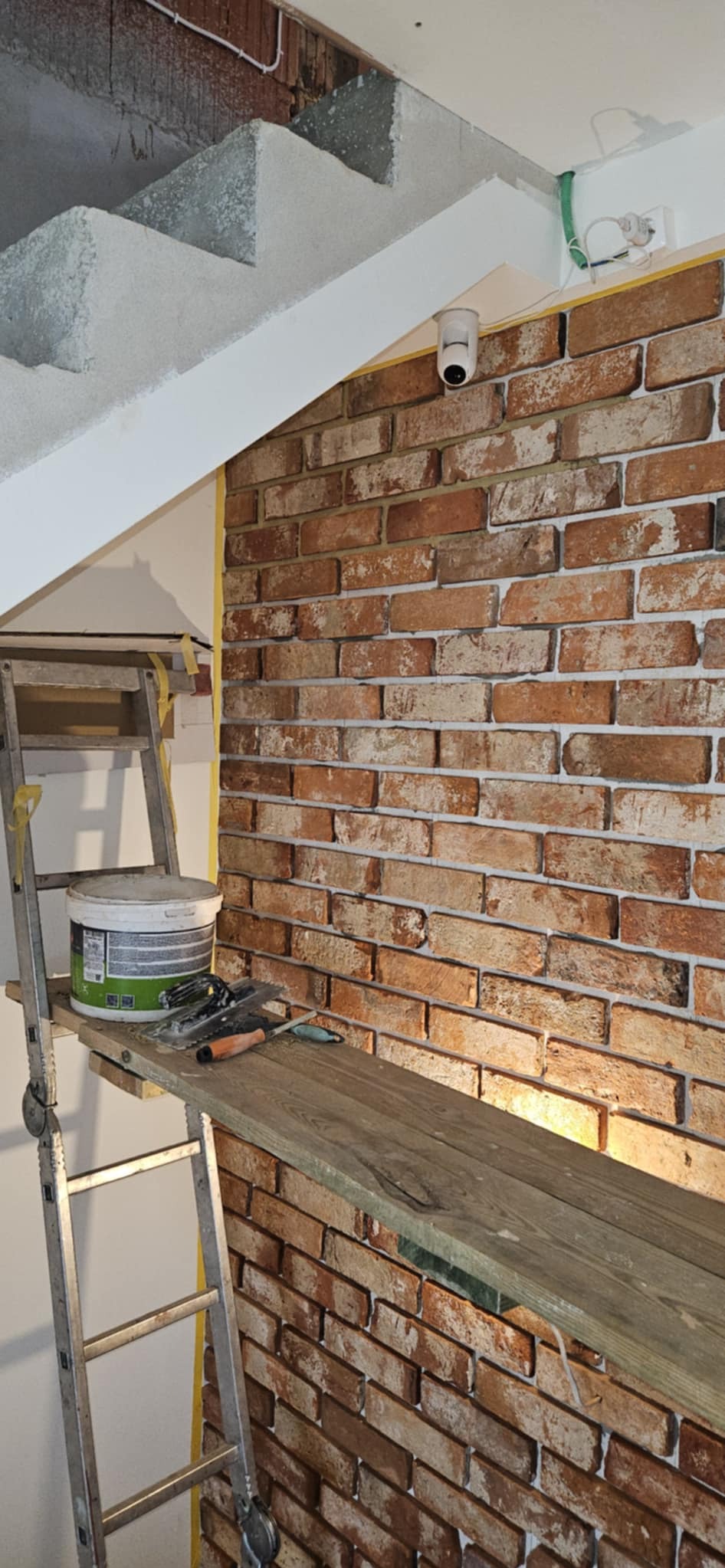 Ściana z cegły klinkierowej w trakcie wykańczania, widoczna drabina z farbą i narzędziami, taśma malarska, oświetlenie sufitowe i kamera monitoringu.
