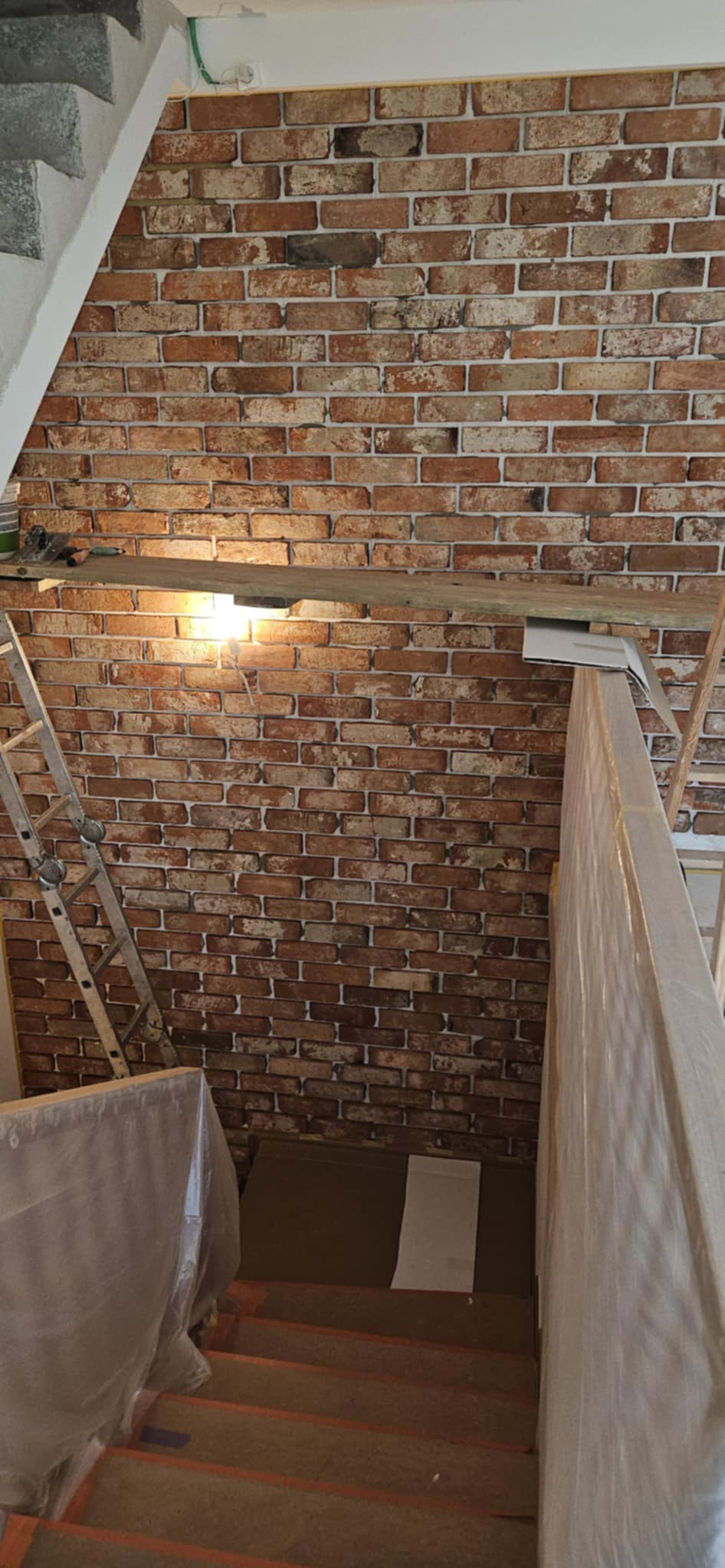 Wykończenie ściany imitacją cegły w klatce schodowej, widoczna drabina i zabezpieczone schody folią malarską. Oświetlenie punktowe na ścianie.