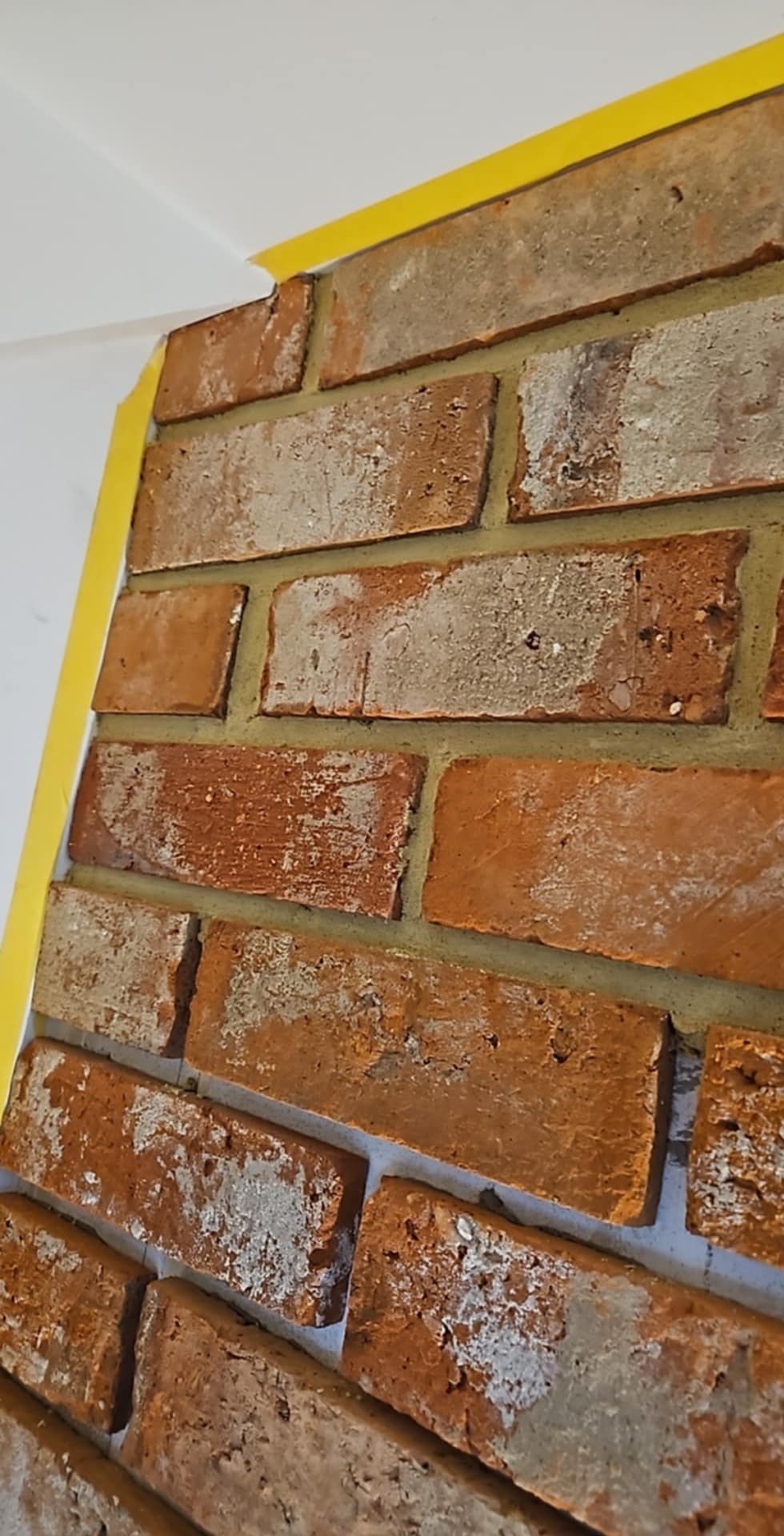 Dekoracyjna ściana z cegły klinkierowej, zabezpieczona żółtą taśmą malarską przy suficie. Widoczna faktura cegieł i fug.