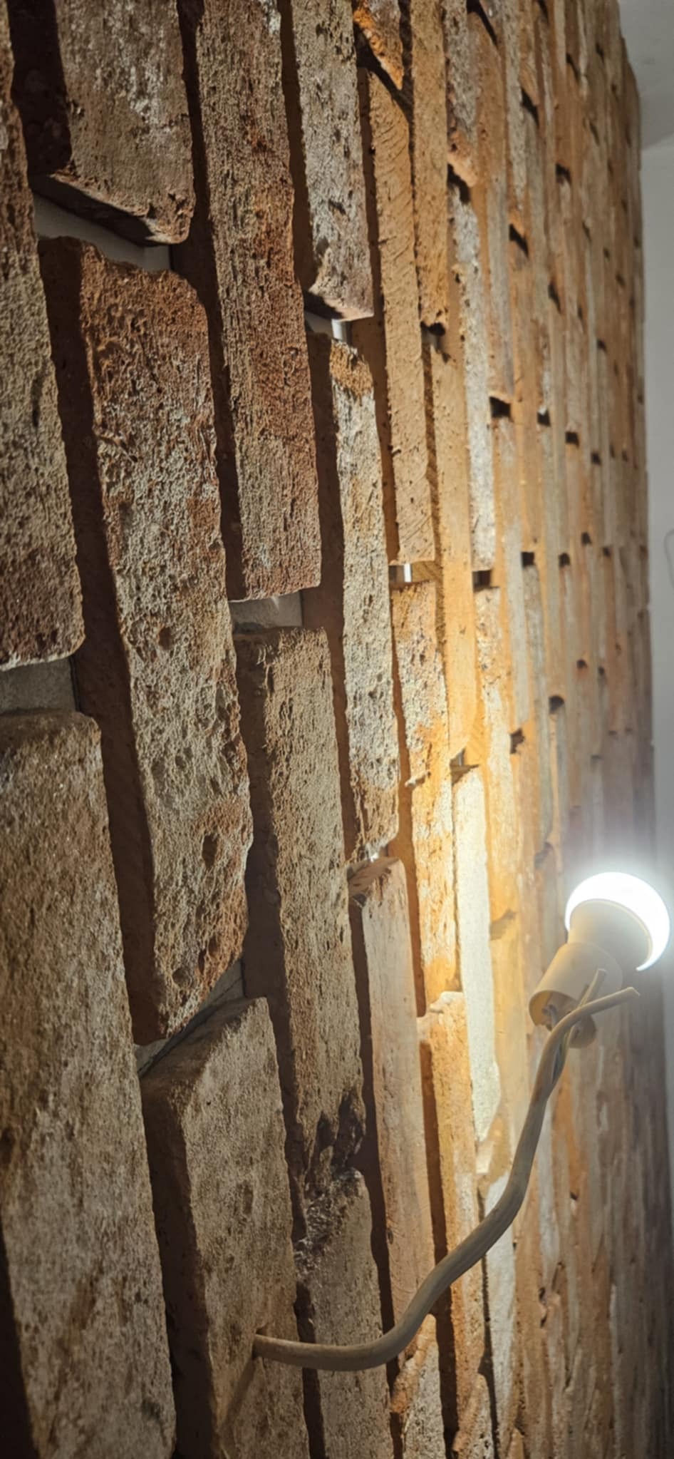 Surowa ściana z cegły z widocznymi spoinami i fragmentem instalacji elektrycznej z żarówką. Detal wnętrza w trakcie remontu lub wykończenia.