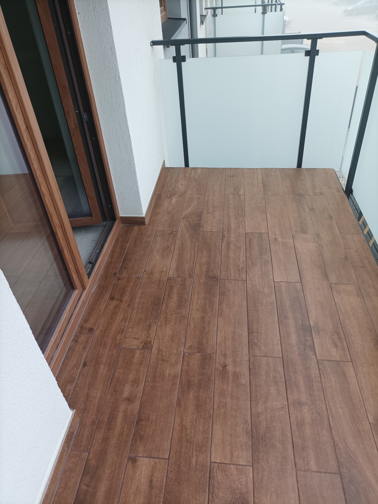 Balkon po remoncie: brązowe płytki imitujące drewno, białe ściany, przesuwne drzwi balkonowe z drewnianą ramą i szklana balustrada z czarnymi elementami.