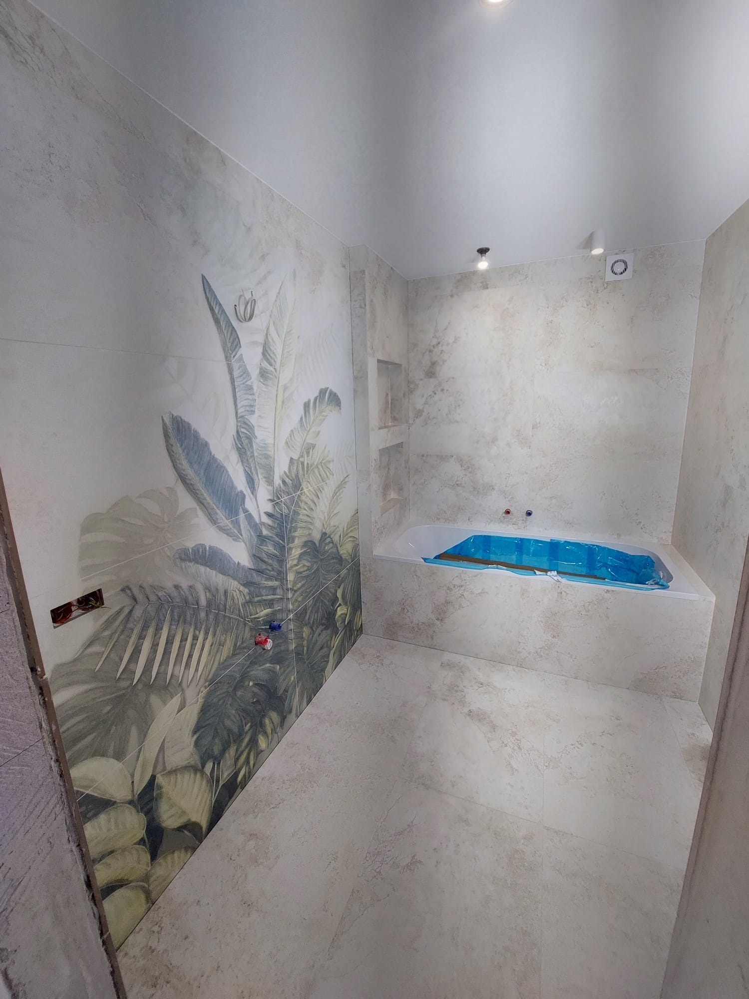 Wykończona łazienka z wanną zabezpieczoną folią. Na ścianie płytki z motywem roślinnym, widoczne punkty elektryczne i armatura. Jasne płytki na podłodze i ścianach.