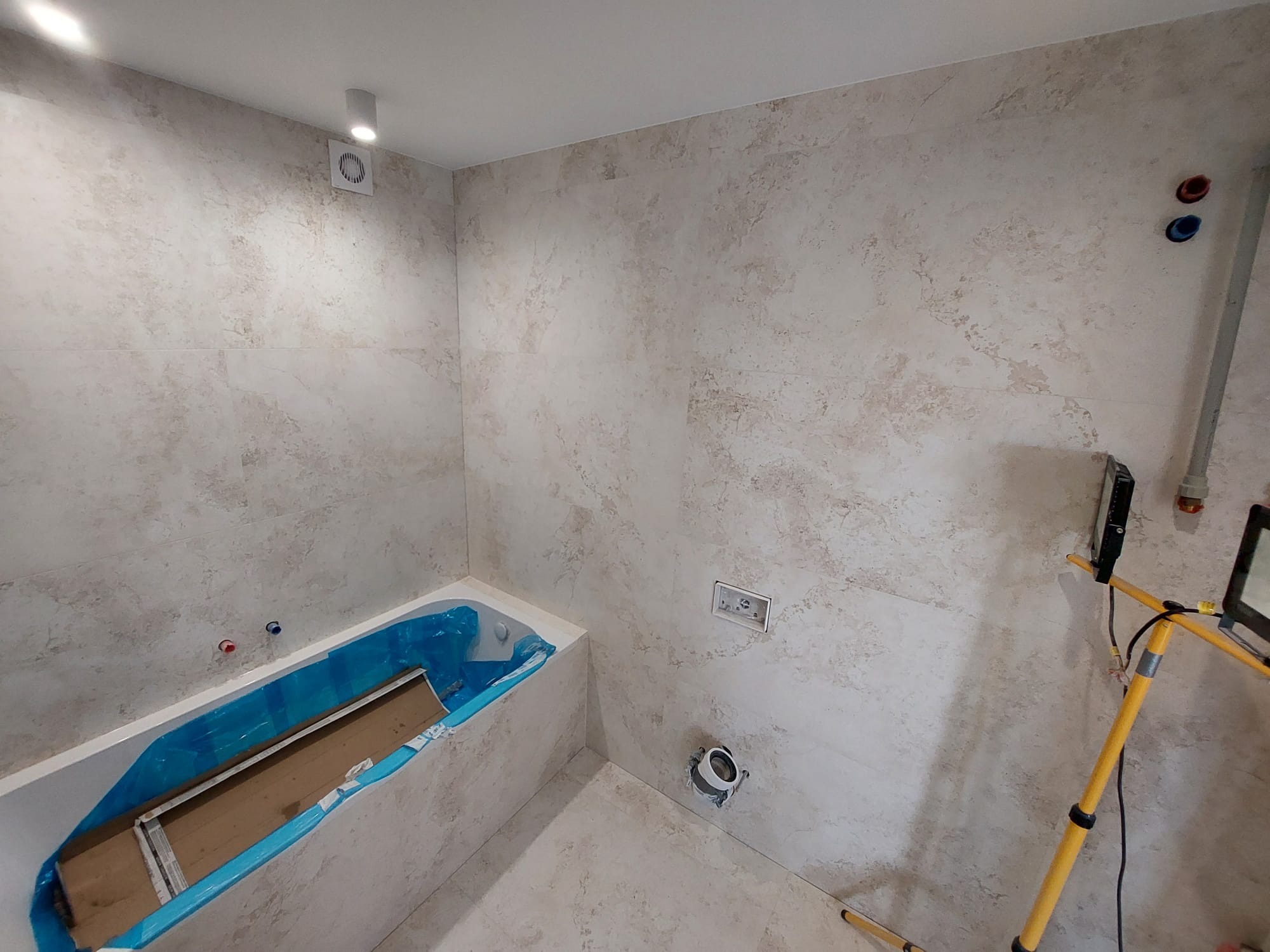 Wykończona łazienka w trakcie montażu. Widoczna wanna zabezpieczona folią, płytki imitujące kamień na ścianach i podłodze. Elementy instalacji wodnej i oświetlenie robocze.