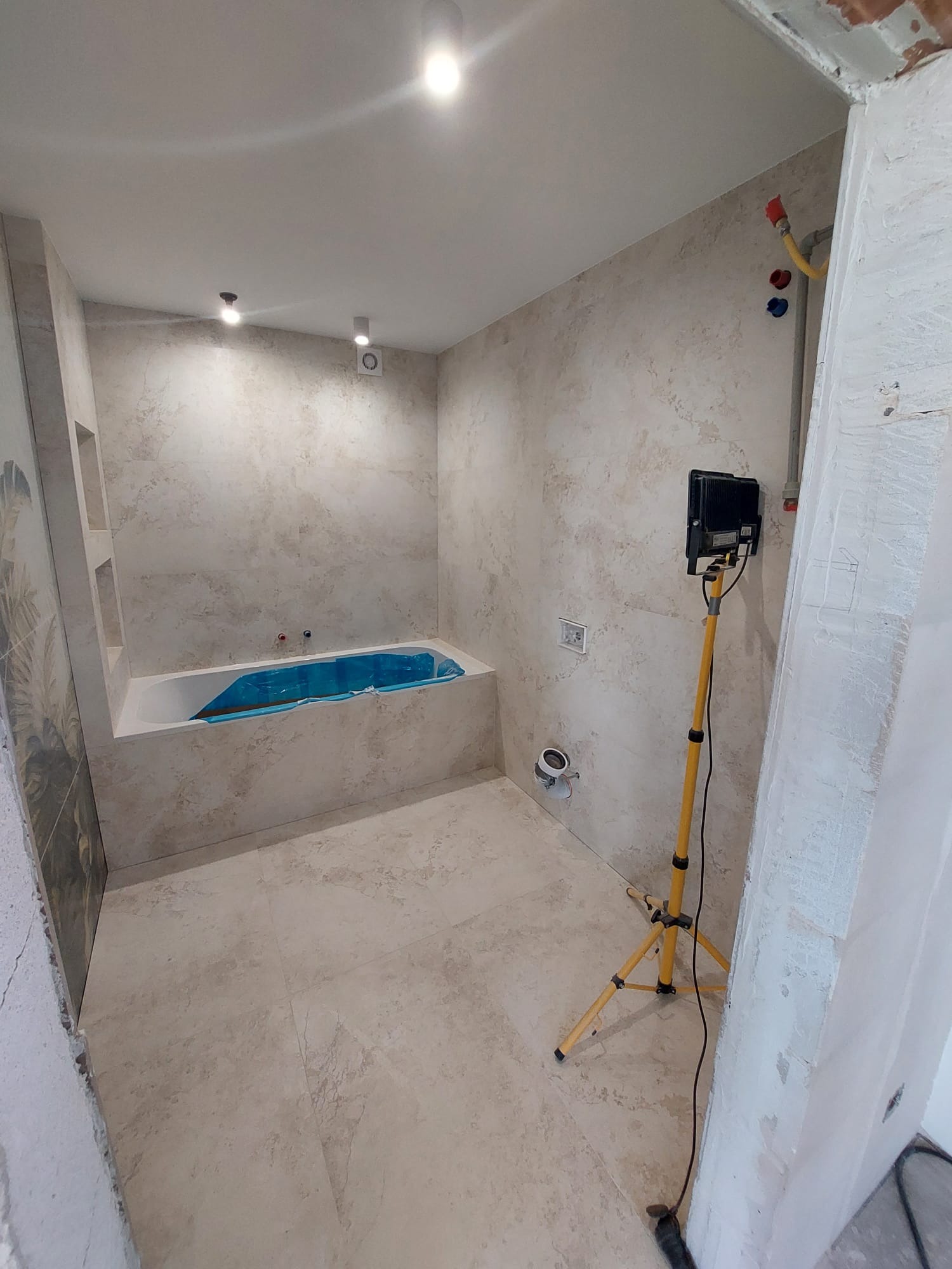 Wykończona łazienka w beżowych płytkach z wanną zabezpieczoną folią, oświetlona lampą budowlaną. Widoczne instalacje i otwarta przestrzeń.