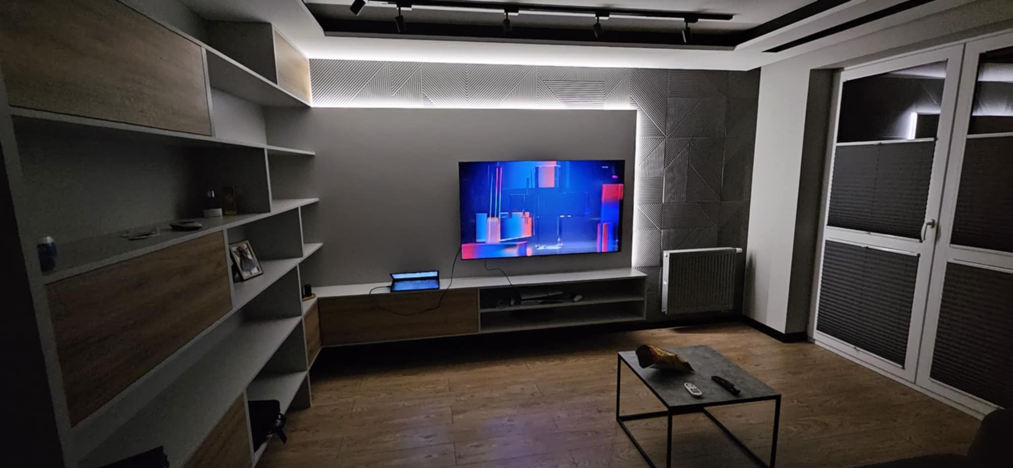 Nowoczesny salon z podświetlaną ścianą 3D za telewizorem, zabudową meblową z półkami i drewnianymi akcentami, oraz stolikiem kawowym na drewnianej podłodze.