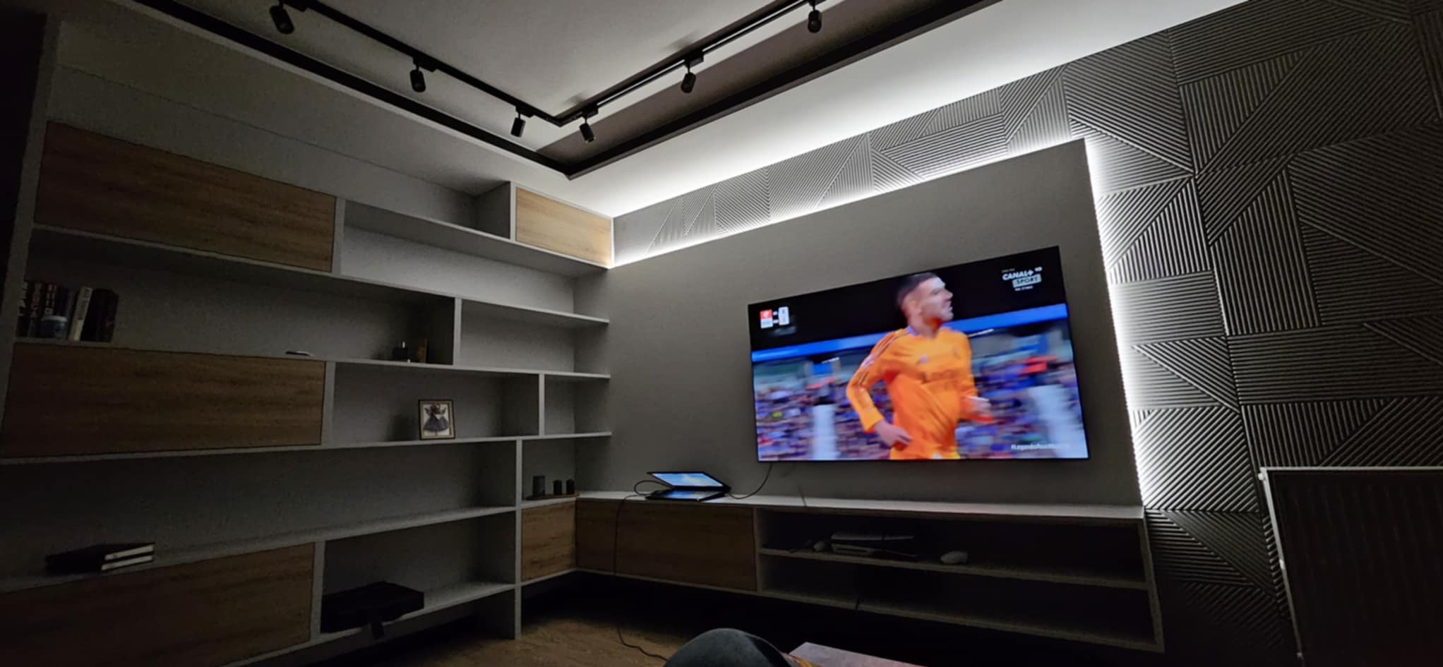 Nowoczesny salon z podświetlaną ścianą dekoracyjną, zabudową RTV i regałem. Na ścianie wisi telewizor z włączonym kanałem sportowym. Oświetlenie punktowe na suficie.