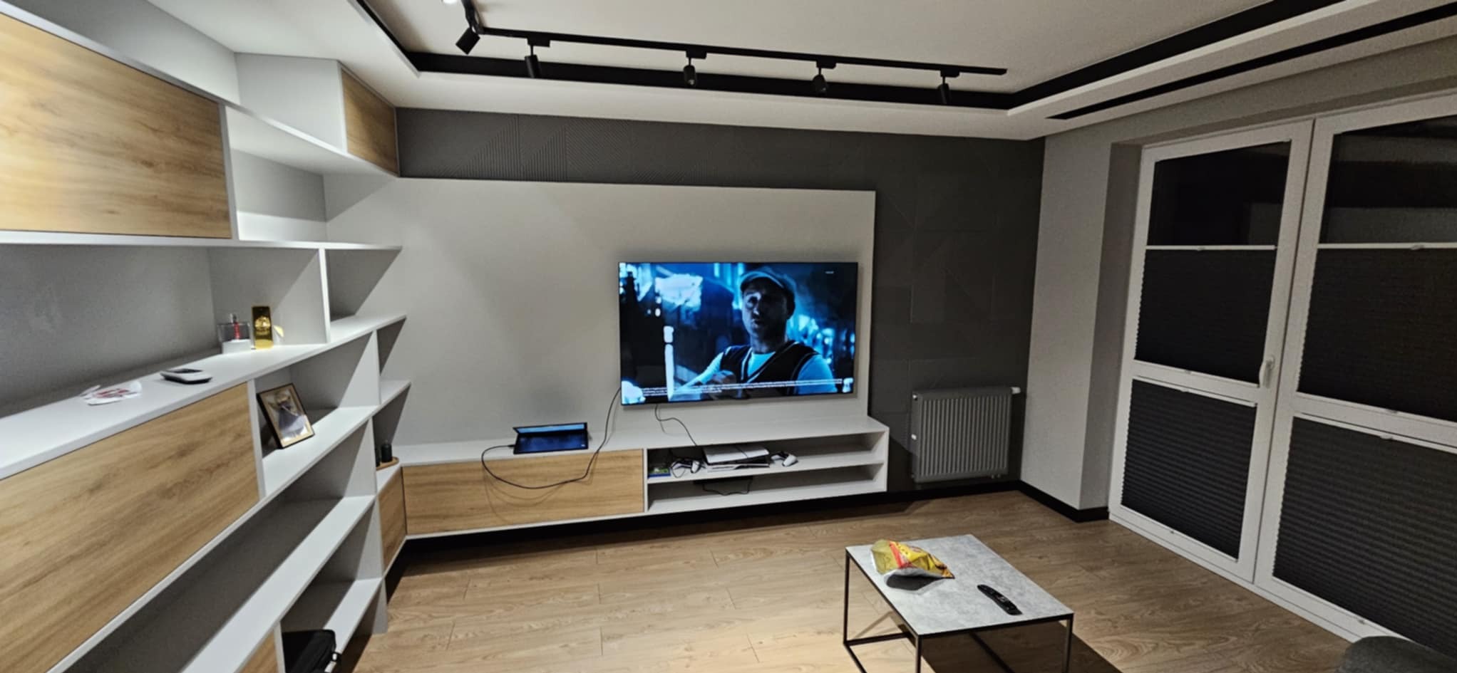 Nowoczesny salon z drewnianą podłogą, biało-szarą zabudową meblową i telewizorem. Widoczne okno z roletami i stolik kawowy z przekąskami.