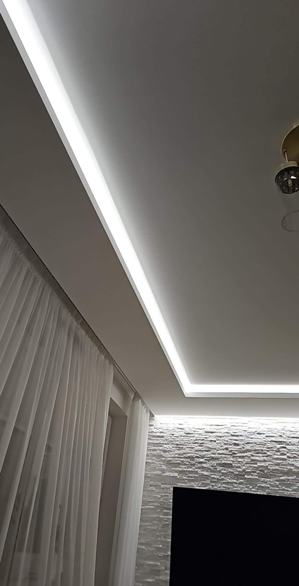 Nowoczesne oświetlenie sufitowe LED w salonie z dekoracyjną ścianą z kamienia i zwiewnymi, białymi zasłonami. Widok z dołu, podkreślający design.