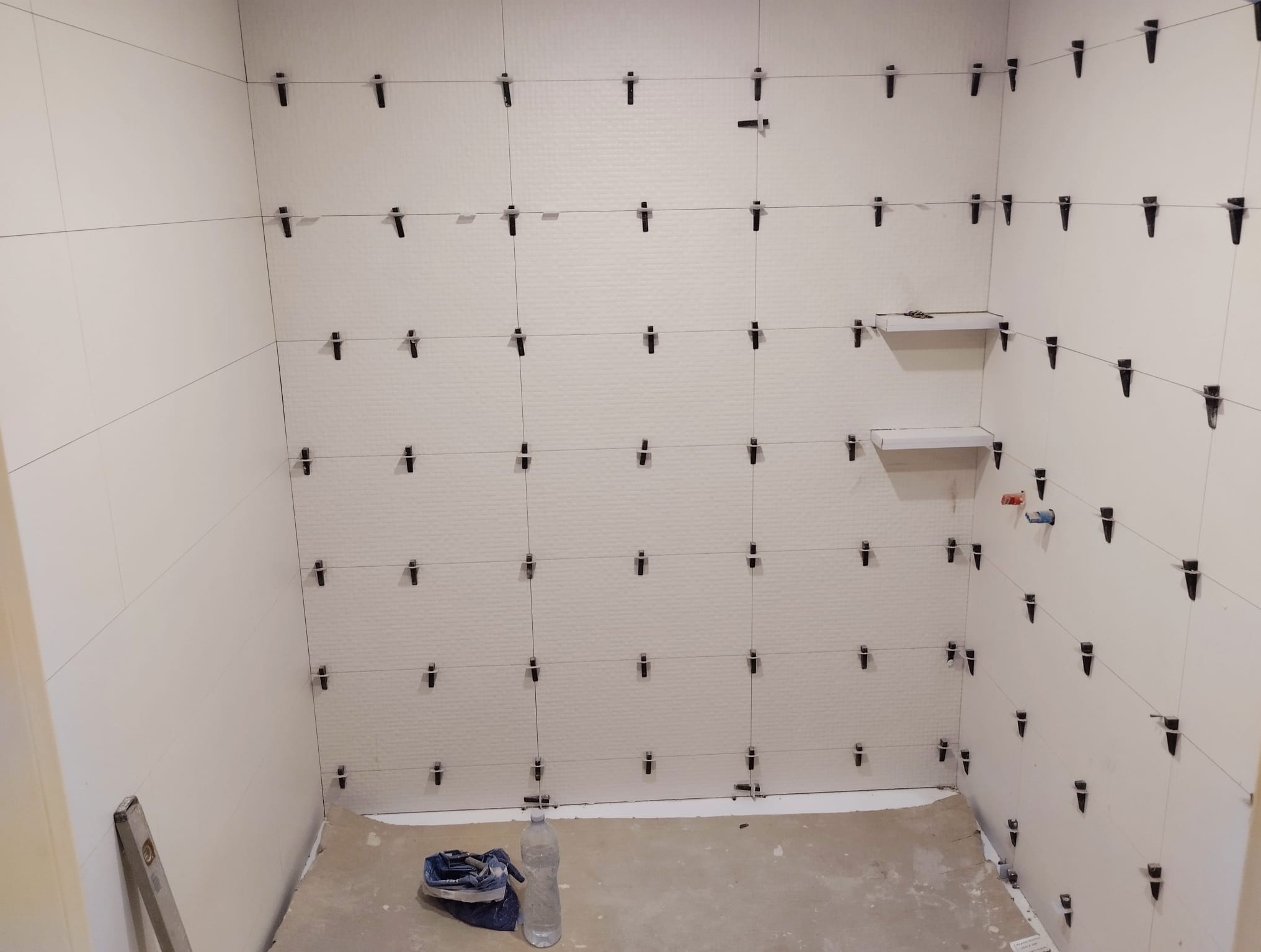Białe płytki ścienne w trakcie montażu w łazience, z użyciem klipsów poziomujących. Widoczne półki i fragment instalacji wodnej. Na podłodze narzędzia i butelka z wodą.