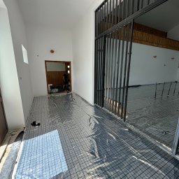 „HESTIA” Instalacje Sanitarne Jakub Rudzewicz - Rozłożona siatka z folią na podłodze, przygotowana do montażu ogrzewania podłogowego w jasnym pomieszczeniu z widoczną konstrukcją metalową.