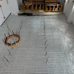 Widok z góry na rozłożone rury ogrzewania podłogowego na srebrnej folii w jasnym wnętrzu. W tle ławki kościelne i wiszący żyrandol.