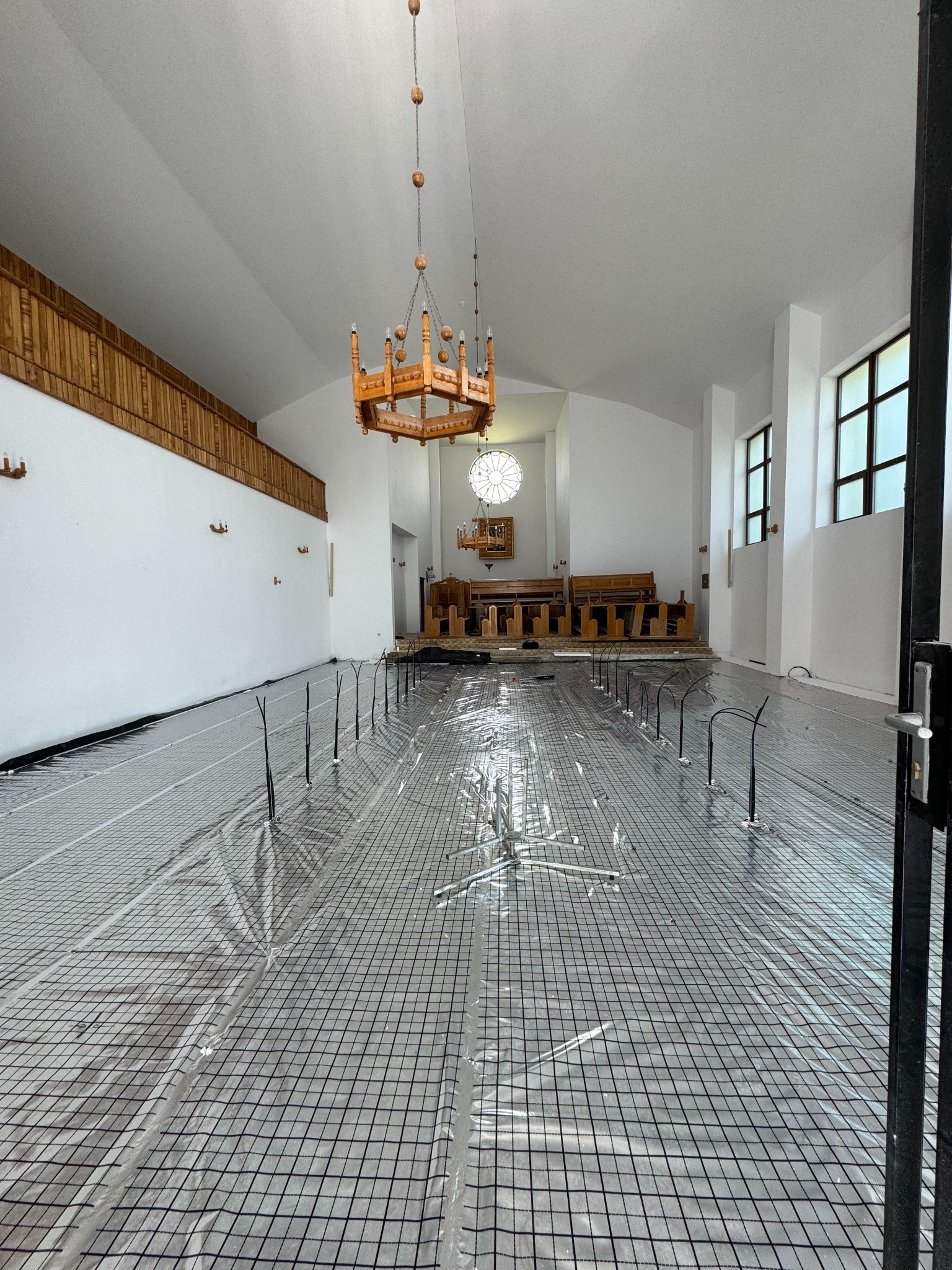 Montaż ogrzewania podłogowego w jasnym wnętrzu z drewnianym żyrandolem. Widoczna siatka grzewcza z wystającymi przewodami, przygotowana do zalania posadzką.