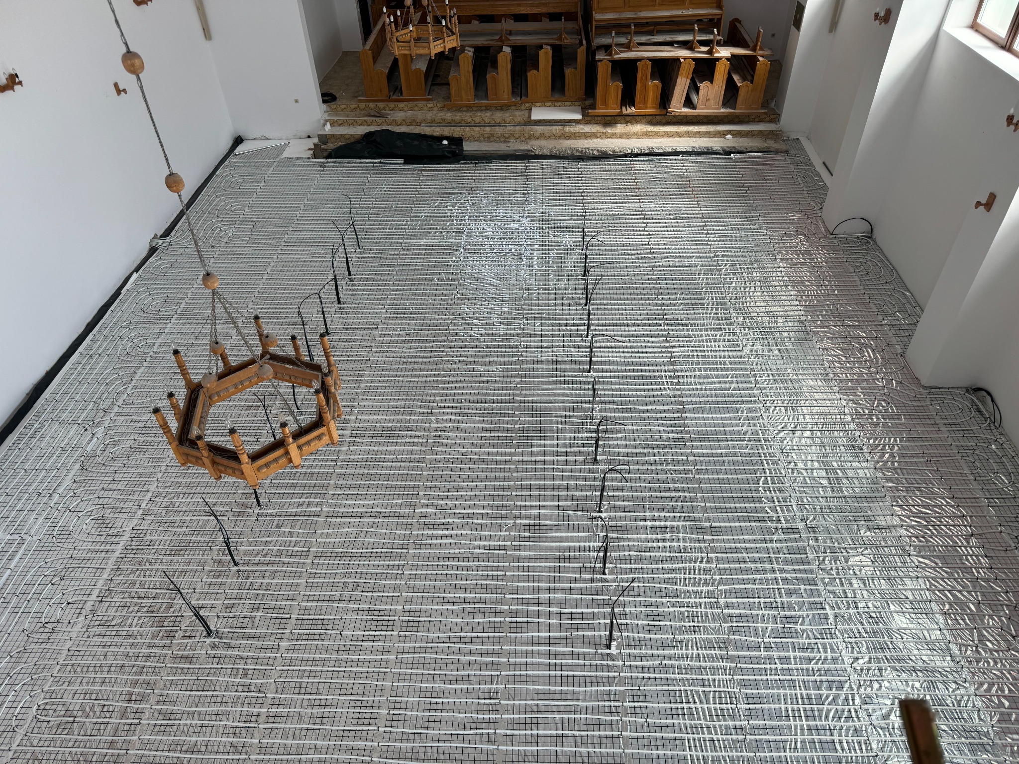 Widok z góry na rozłożone rury ogrzewania podłogowego na srebrnej folii w jasnym wnętrzu. W tle ławki kościelne i wiszący żyrandol.