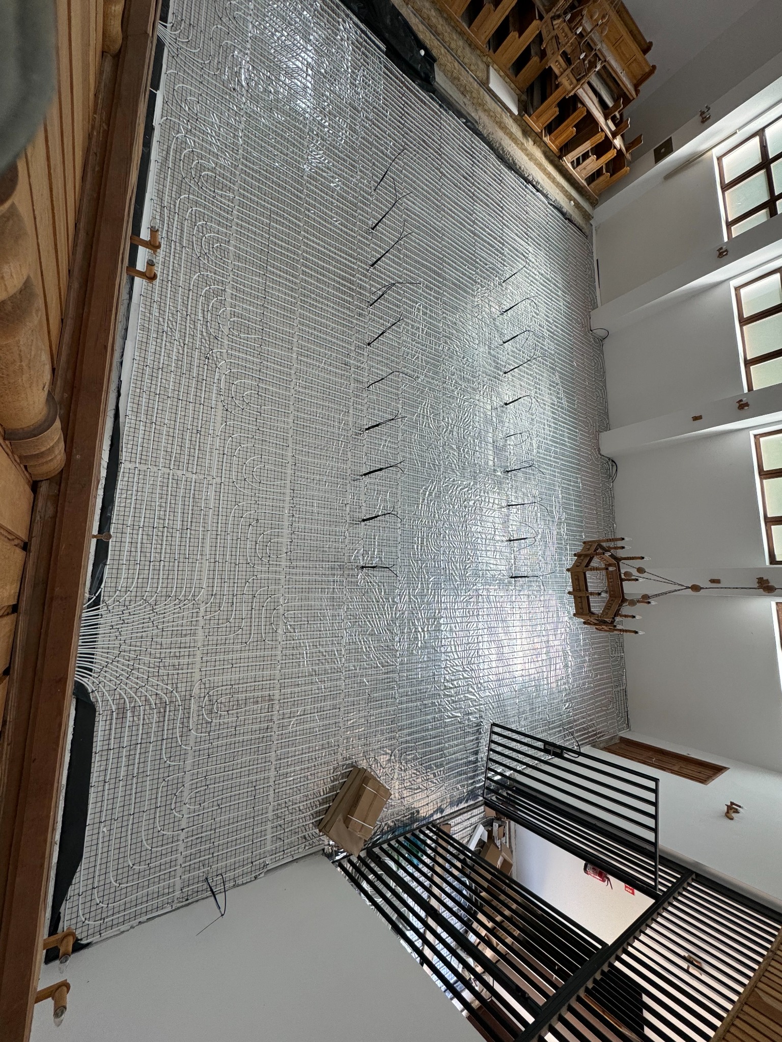 Montaż ogrzewania podłogowego w budynku z drewnianymi elementami wykończeniowymi. Widoczne białe rury na srebrnej folii izolacyjnej i fragment drewnianego żyrandola.