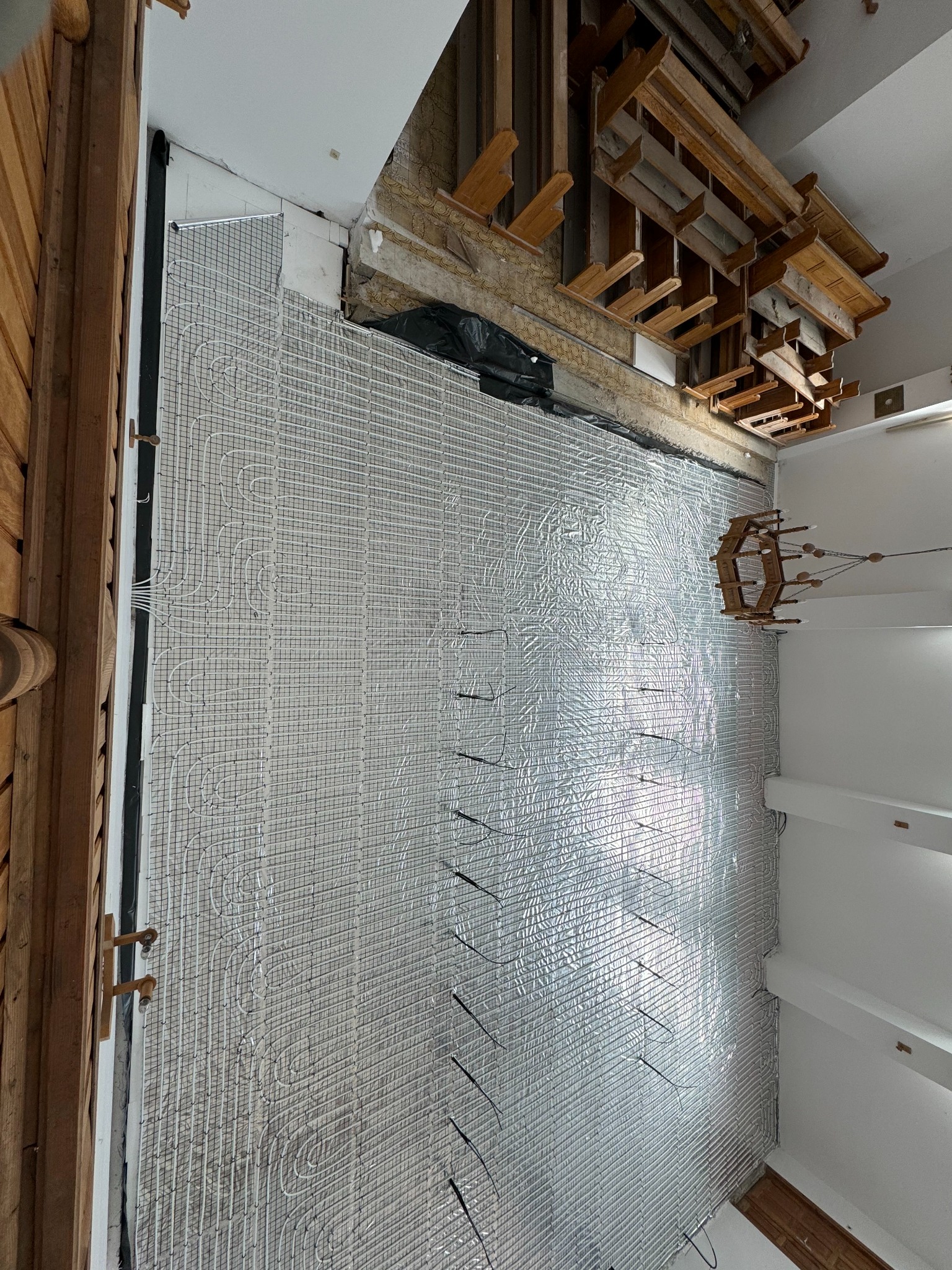 Instalacja ogrzewania podłogowego: srebrna mata grzewcza z rurkami, widok z góry, surowy stan wykończenia, drewniane elementy konstrukcyjne w tle.