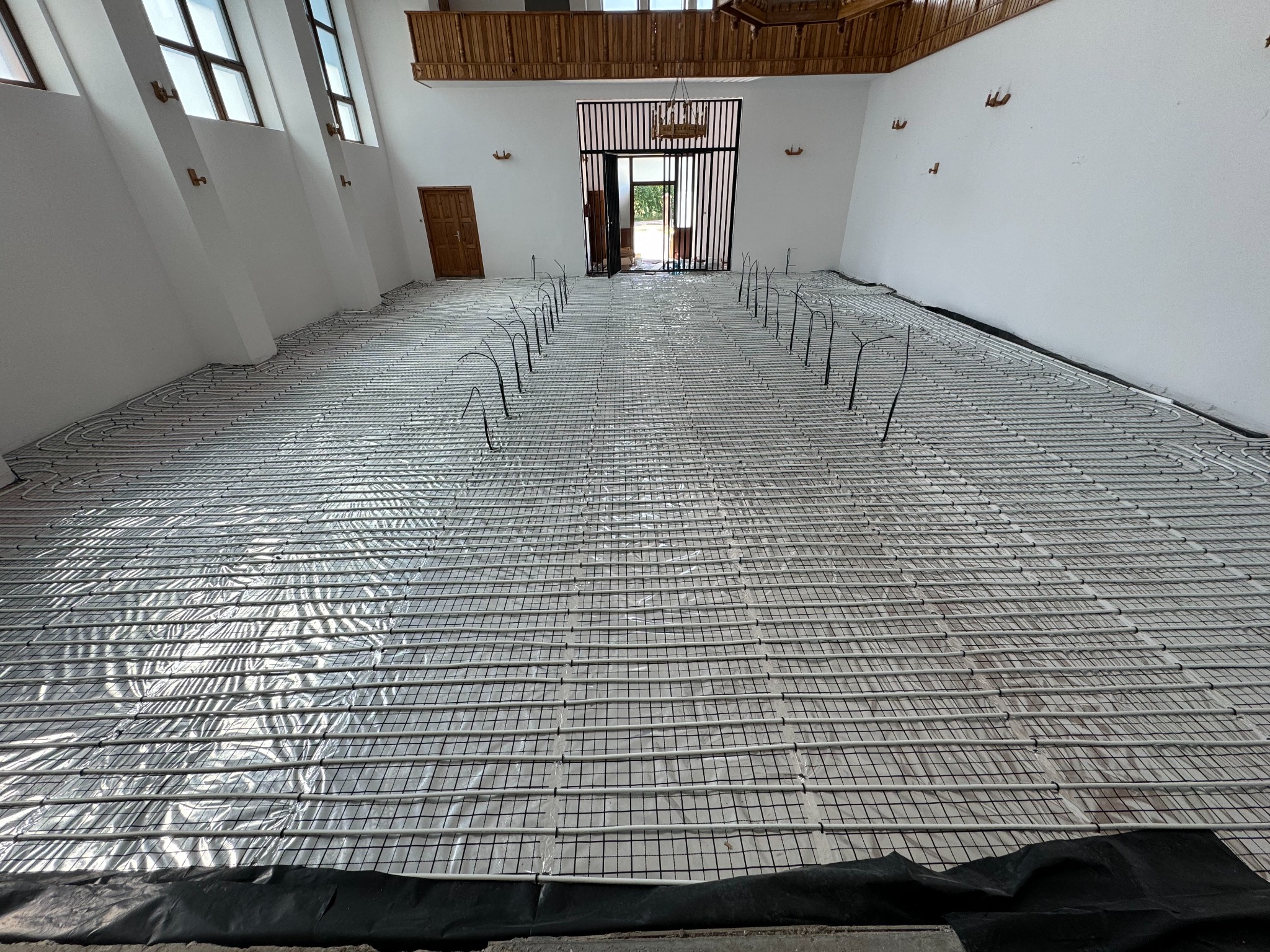 Białe rury ogrzewania podłogowego ułożone na folii w przestronnym wnętrzu z drewnianą antresolą i oknami w skośnej ścianie. Widok na rozłożoną instalację przed zalaniem posadzką.