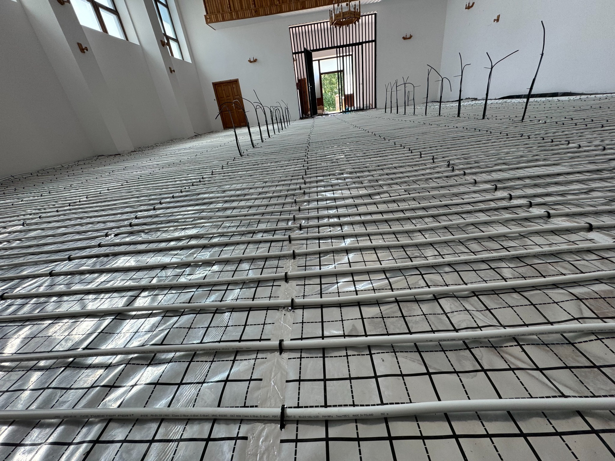 Instalacja ogrzewania podłogowego w przestronnym wnętrzu z białymi ścianami i dużymi oknami. Równoległe rury na folii z siatką, pionowe wyprowadzenia rur.
