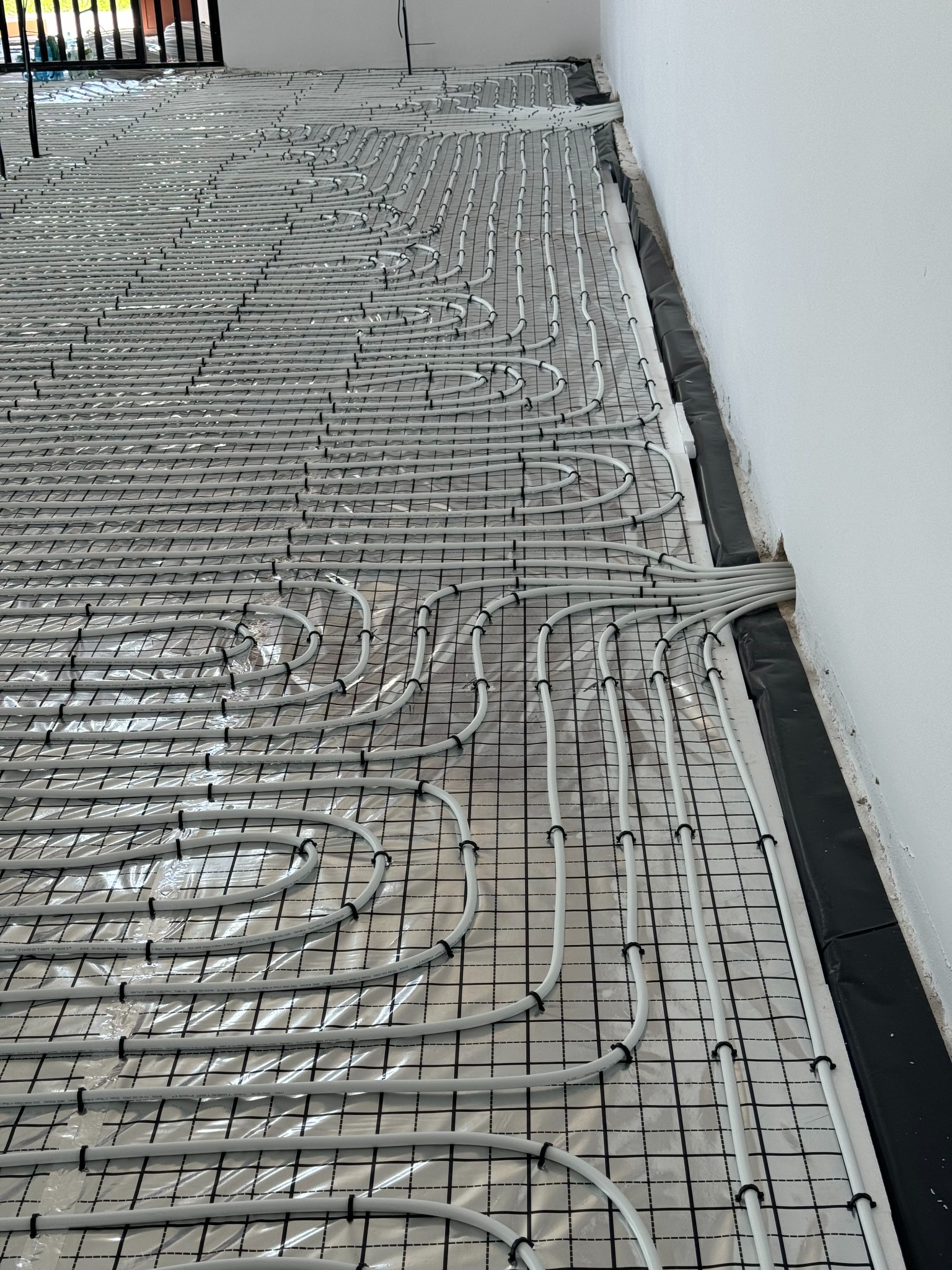 Ułożone, białe rury ogrzewania podłogowego na folii izolacyjnej z czarną siatką, przymocowane czarnymi uchwytami, widok z góry, przy białej ścianie.