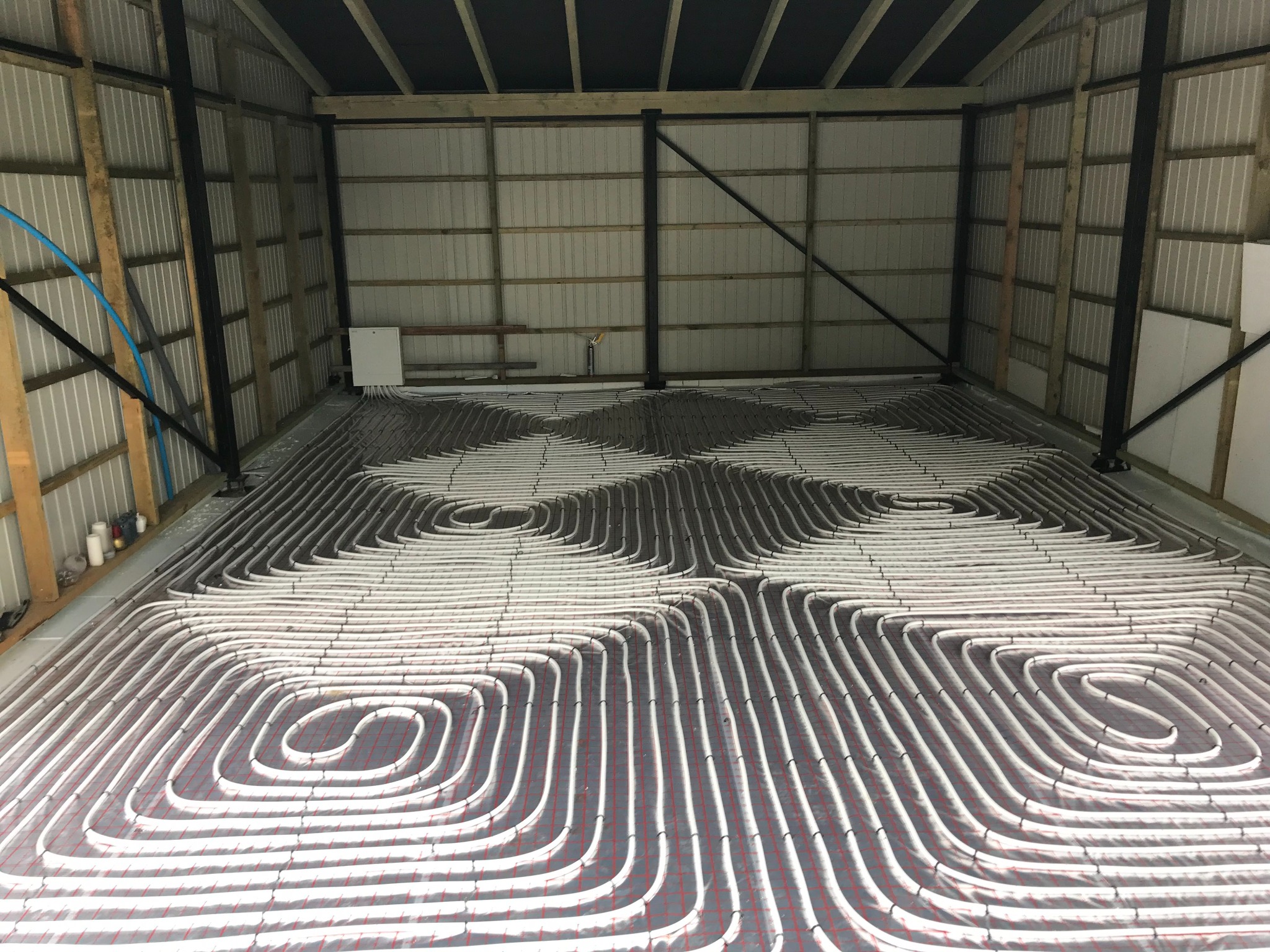 Ogrzewanie podłogowe w hali: białe rury ułożone spiralnie na szarej macie z czerwoną siatką, widoczne elementy konstrukcyjne hali.
