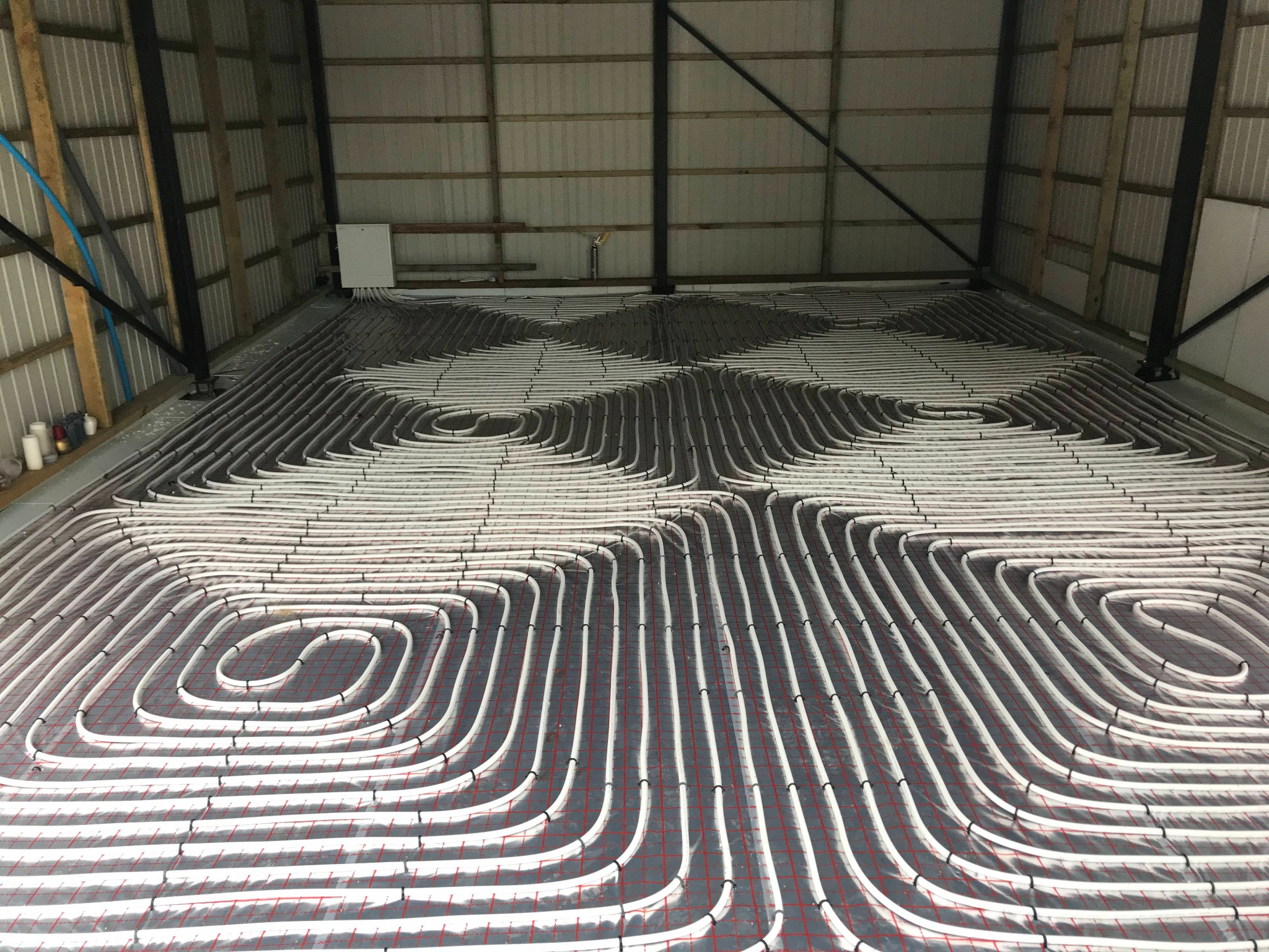 Białe rury ogrzewania podłogowego ułożone spiralnie na folii z czerwoną siatką, w surowym wnętrzu hali z metalową konstrukcją dachu.