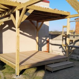 Eagle Tech Krystian Orłowski - Drewniana altana z tarasem, widok z boku, jasne drewno, częściowe zacienienie, elementy konstrukcyjne w tle. Nowoczesna architektura ogrodowa.