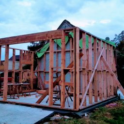 Eagle Tech Krystian Orłowski - Surowy szkielet drewnianej konstrukcji domu w trakcie budowy, widoczne elementy konstrukcyjne i fragment zielonej membrany. Tło stanowią drzewa i zabudowania.