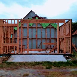 Eagle Tech Krystian Orłowski - Konstrukcja szkieletowa domu w trakcie budowy, widoczne drewniane ramy i zielona membrana. W tle budynek gospodarczy i traktor.