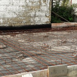 Eagle Tech Krystian Orłowski - Zbrojenie płyty fundamentowej z użyciem dystansów, widoczne szalunki i izolacja. Ujęcie z góry, z widocznym murem w tle.