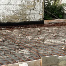 Zbrojenie płyty fundamentowej z użyciem dystansów, widoczne szalunki i izolacja. Ujęcie z góry, z widocznym murem w tle.