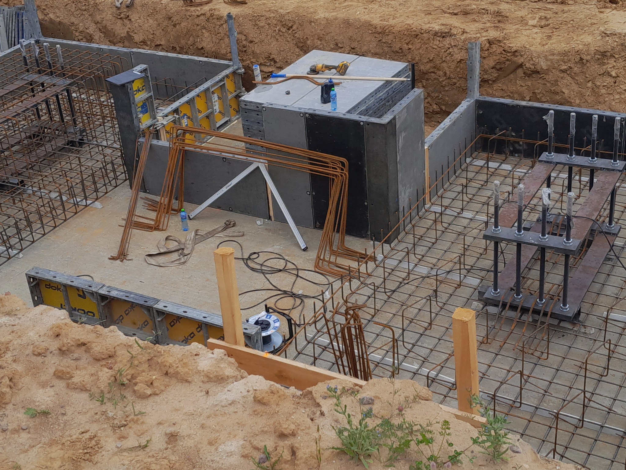 Zbrojenie fundamentów z szalunkami i wystającymi prętami, widok z góry. Na betonowej płycie leżą narzędzia i butelka wody. Wykopane ziemne tło.