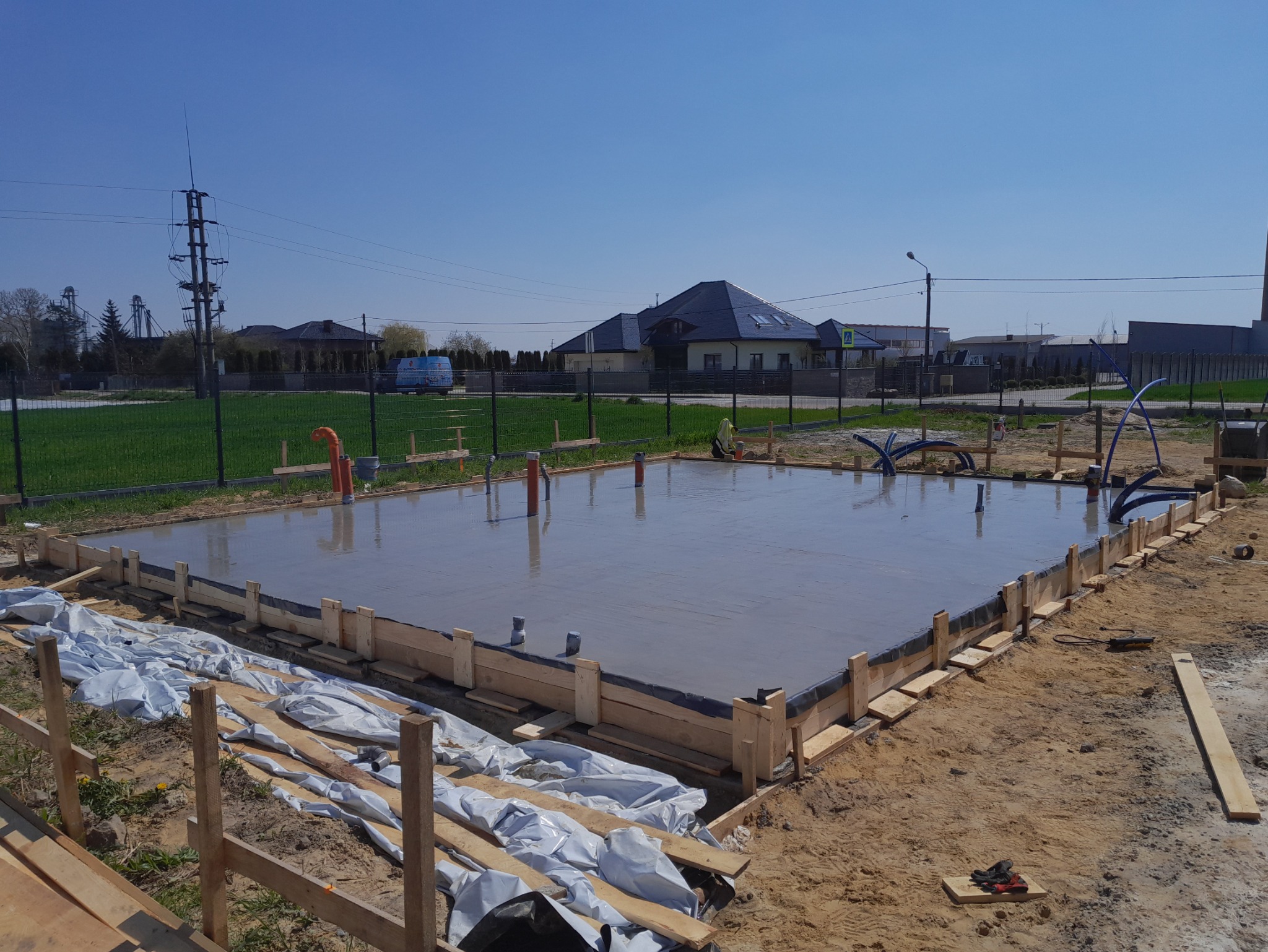 Zalany fundament pod budowę domu, z widocznym szalunkiem i wystającymi rurami. W tle domy jednorodzinne i słup energetyczny.