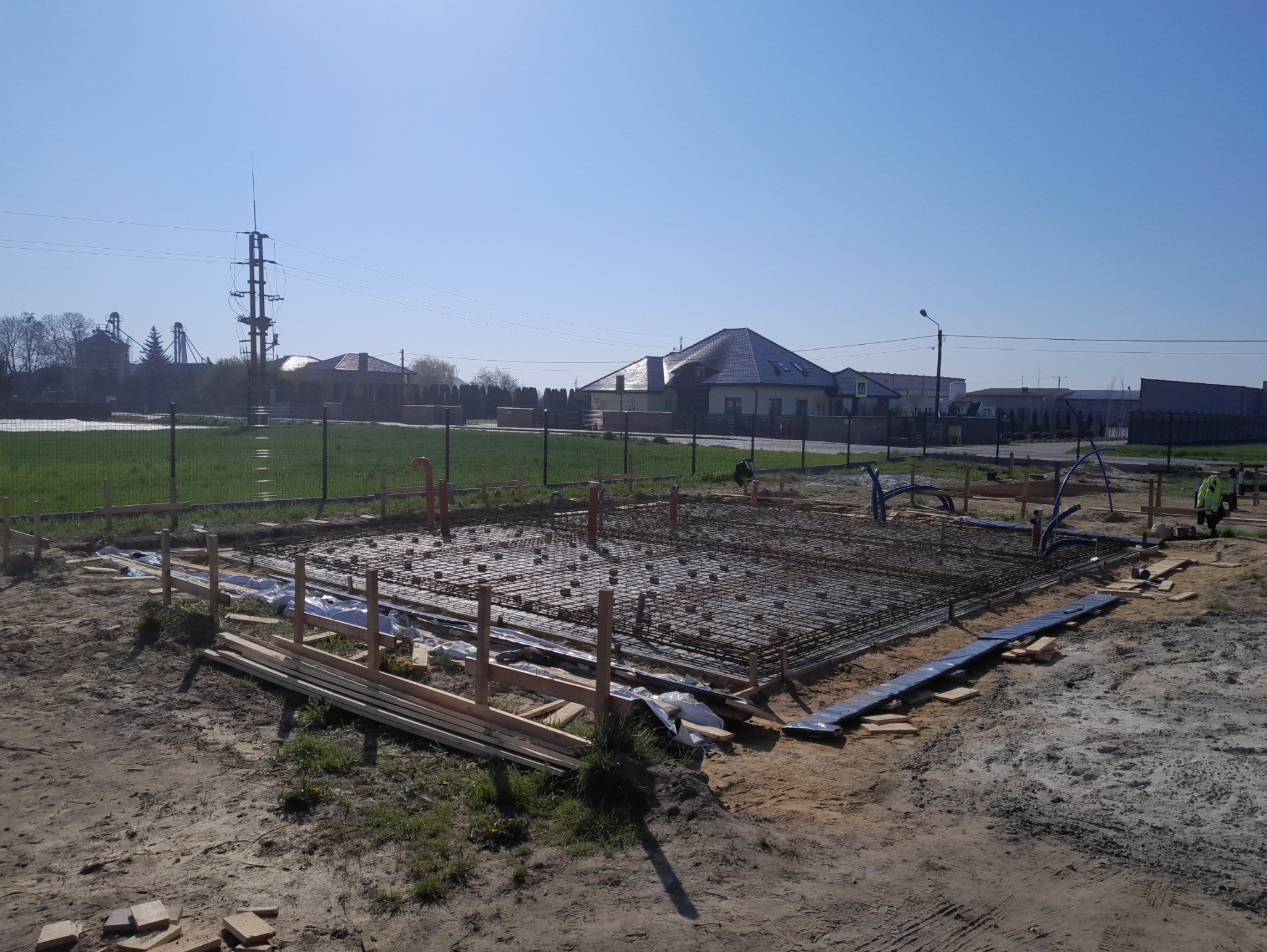 Zbrojenie fundamentu domu jednorodzinnego na działce z widokiem na okoliczną zabudowę i słup wysokiego napięcia w tle, w słoneczny dzień.