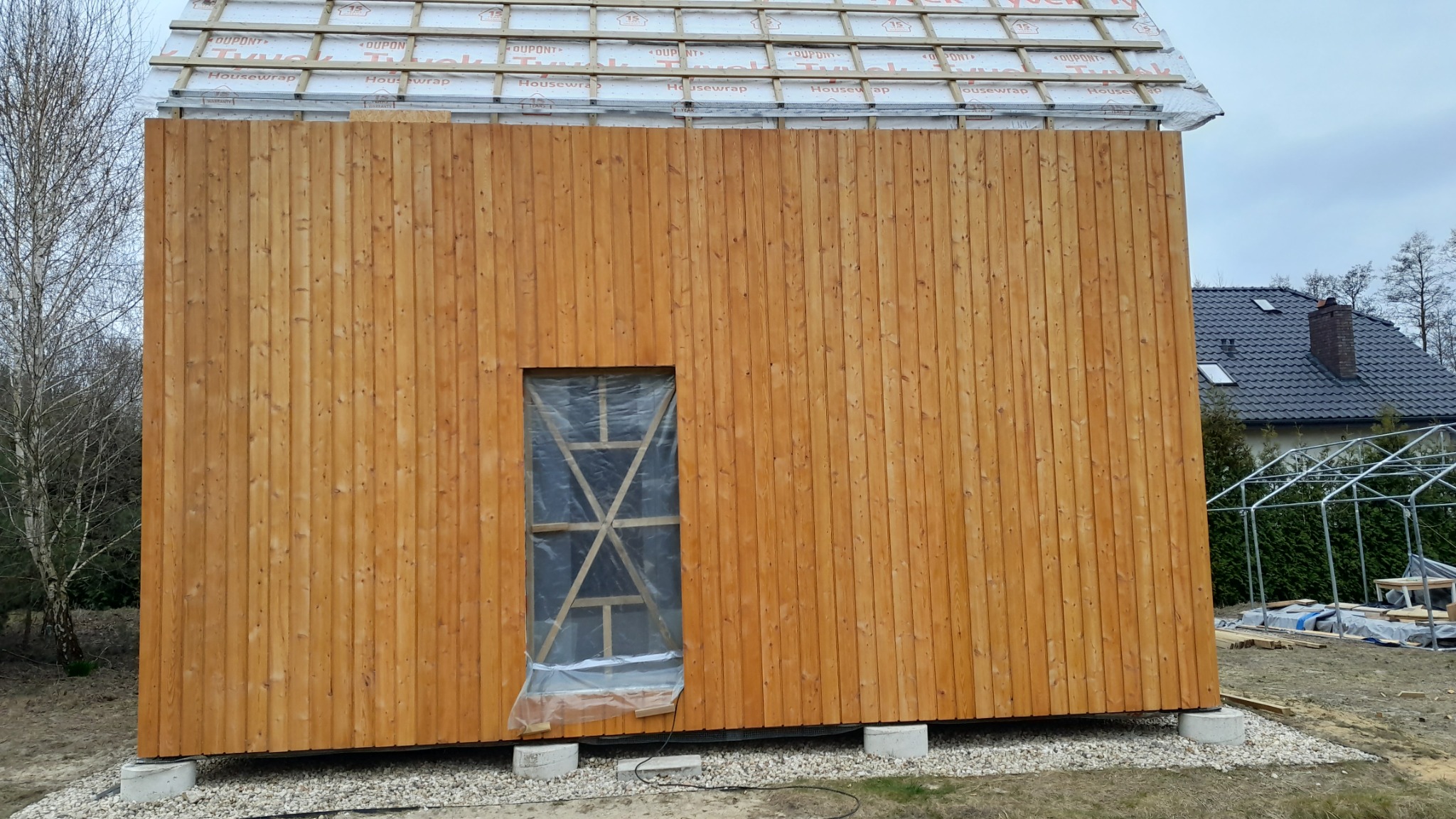 Surowa elewacja domu szkieletowego z pionowych desek, okno zabezpieczone folią i drewnianą konstrukcją, widoczne elementy dachu i fundamentu.