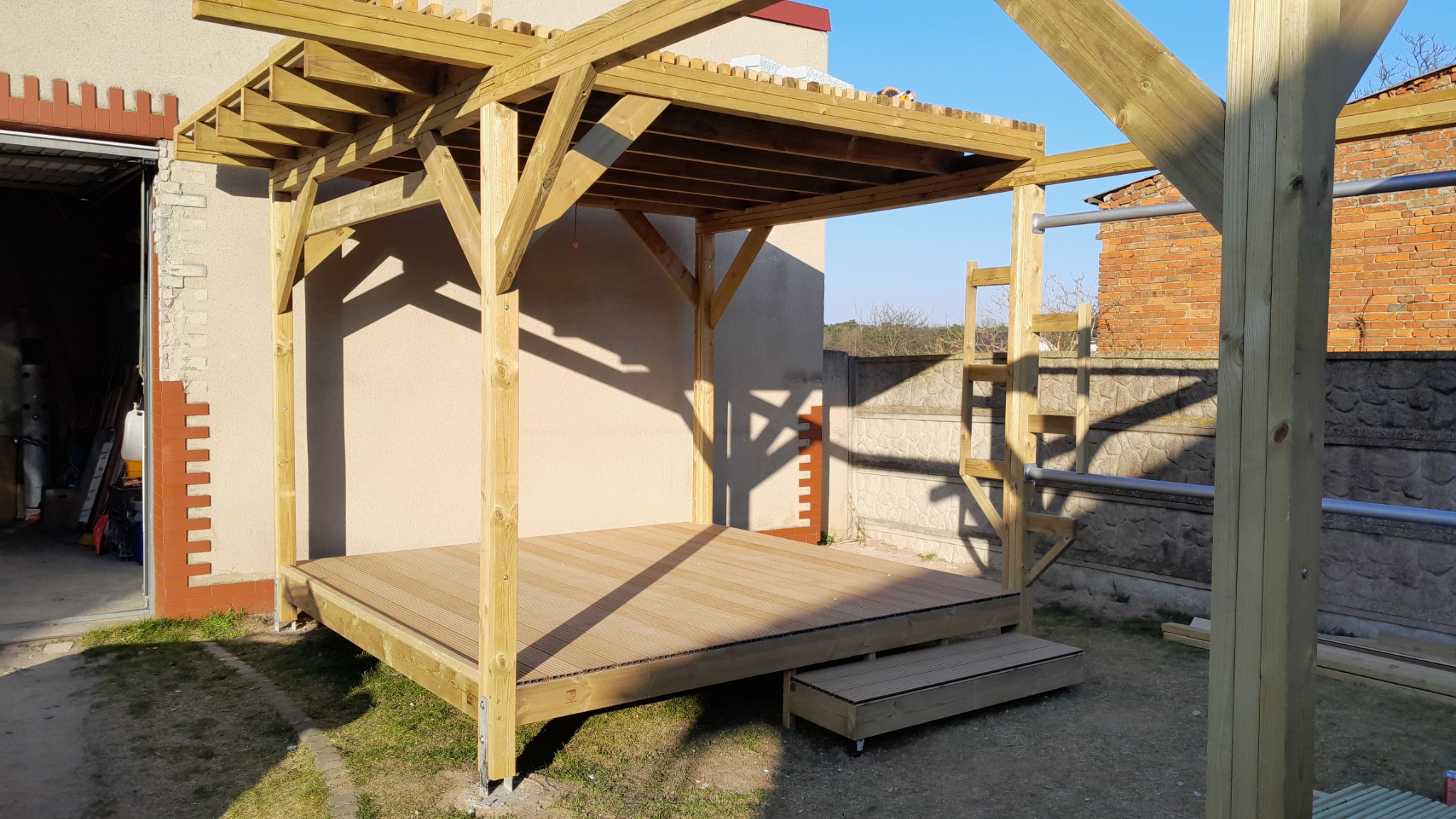 Drewniana altana z tarasem, widok z boku, jasne drewno, częściowe zacienienie, elementy konstrukcyjne w tle. Nowoczesna architektura ogrodowa.