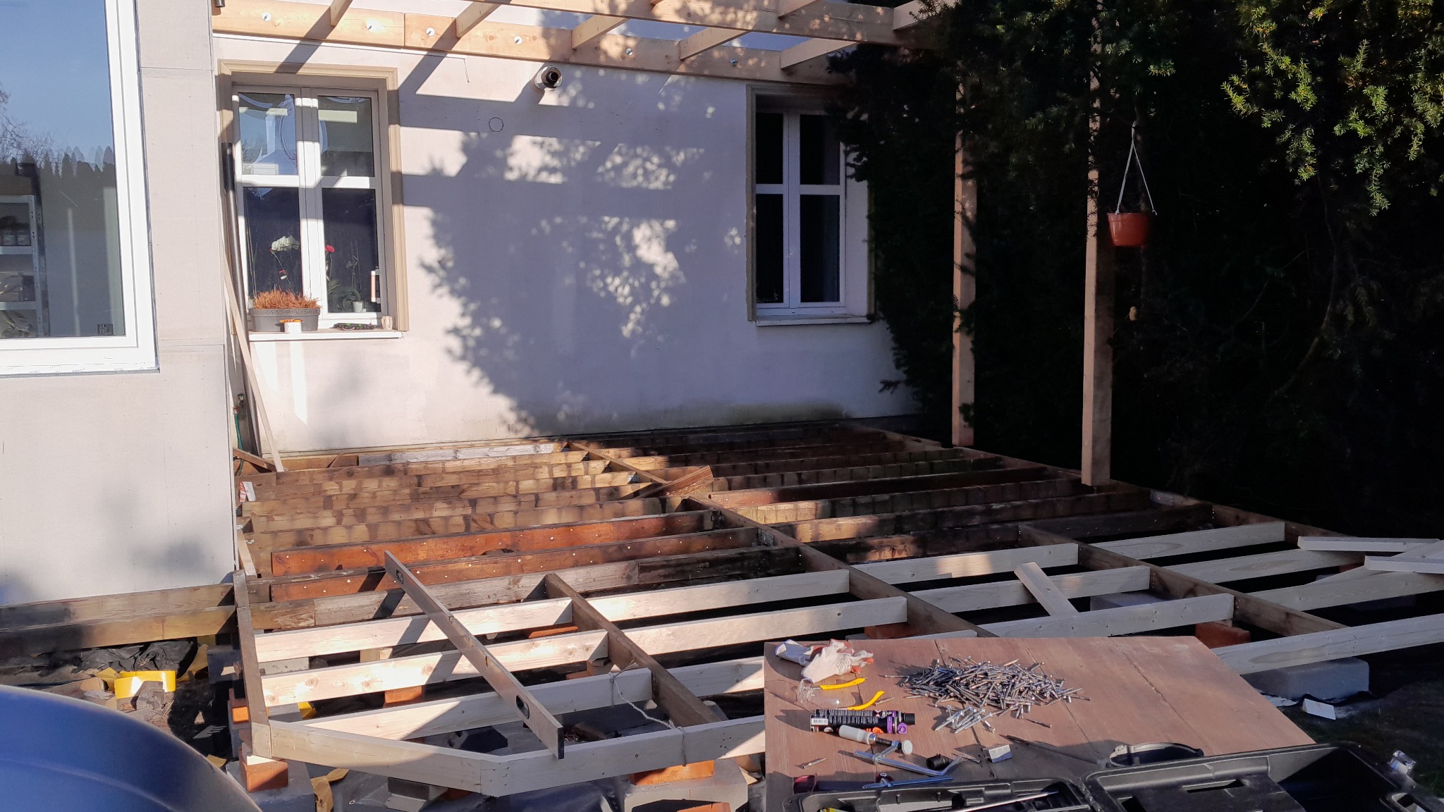 Konstrukcja drewnianego tarasu w trakcie budowy, widoczne legary i narzędzia na desce. W tle biały budynek z oknami i zieleń.