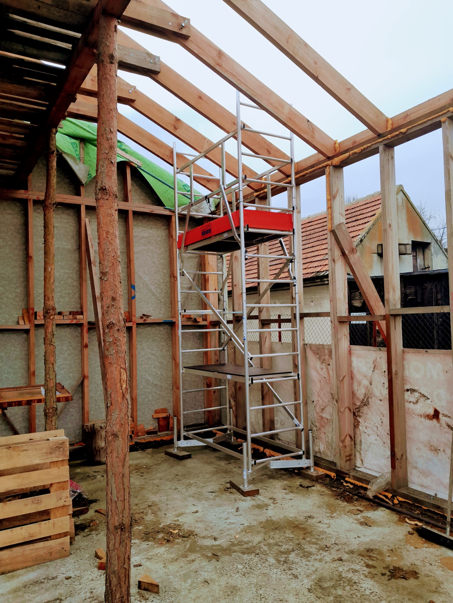 Wnętrze w trakcie budowy: drewniana konstrukcja szkieletowa z izolacją, rusztowanie na środku, widoczne elementy mocowań i pianka uszczelniająca.