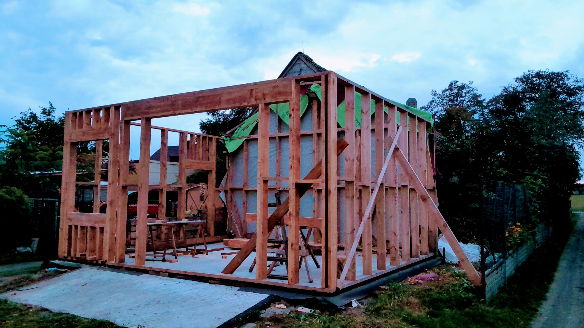 Surowy szkielet drewnianej konstrukcji domu w trakcie budowy, widoczne elementy konstrukcyjne i fragment zielonej membrany. Tło stanowią drzewa i zabudowania.