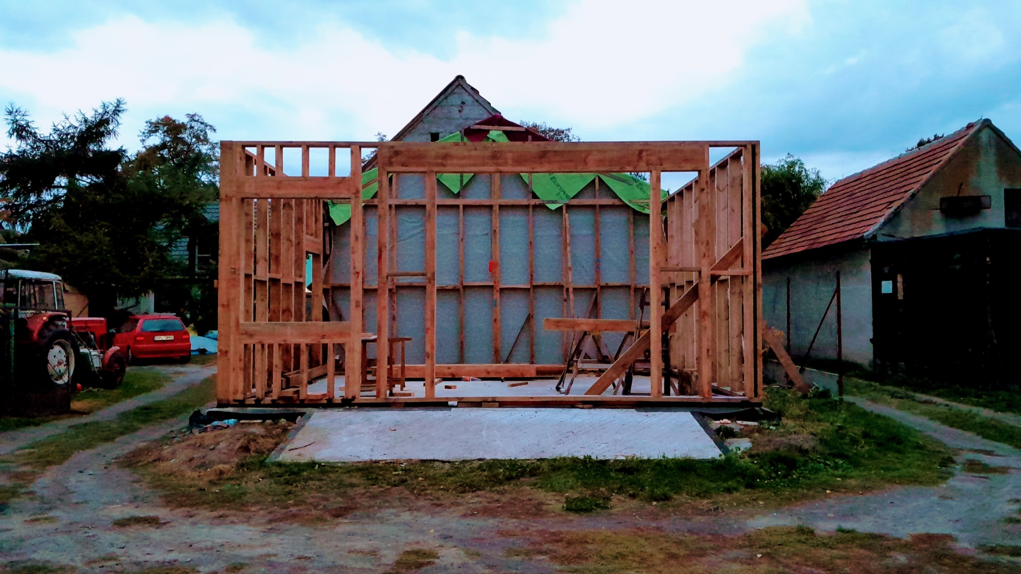 Konstrukcja szkieletowa domu w trakcie budowy, widoczne drewniane ramy i zielona membrana. W tle budynek gospodarczy i traktor.