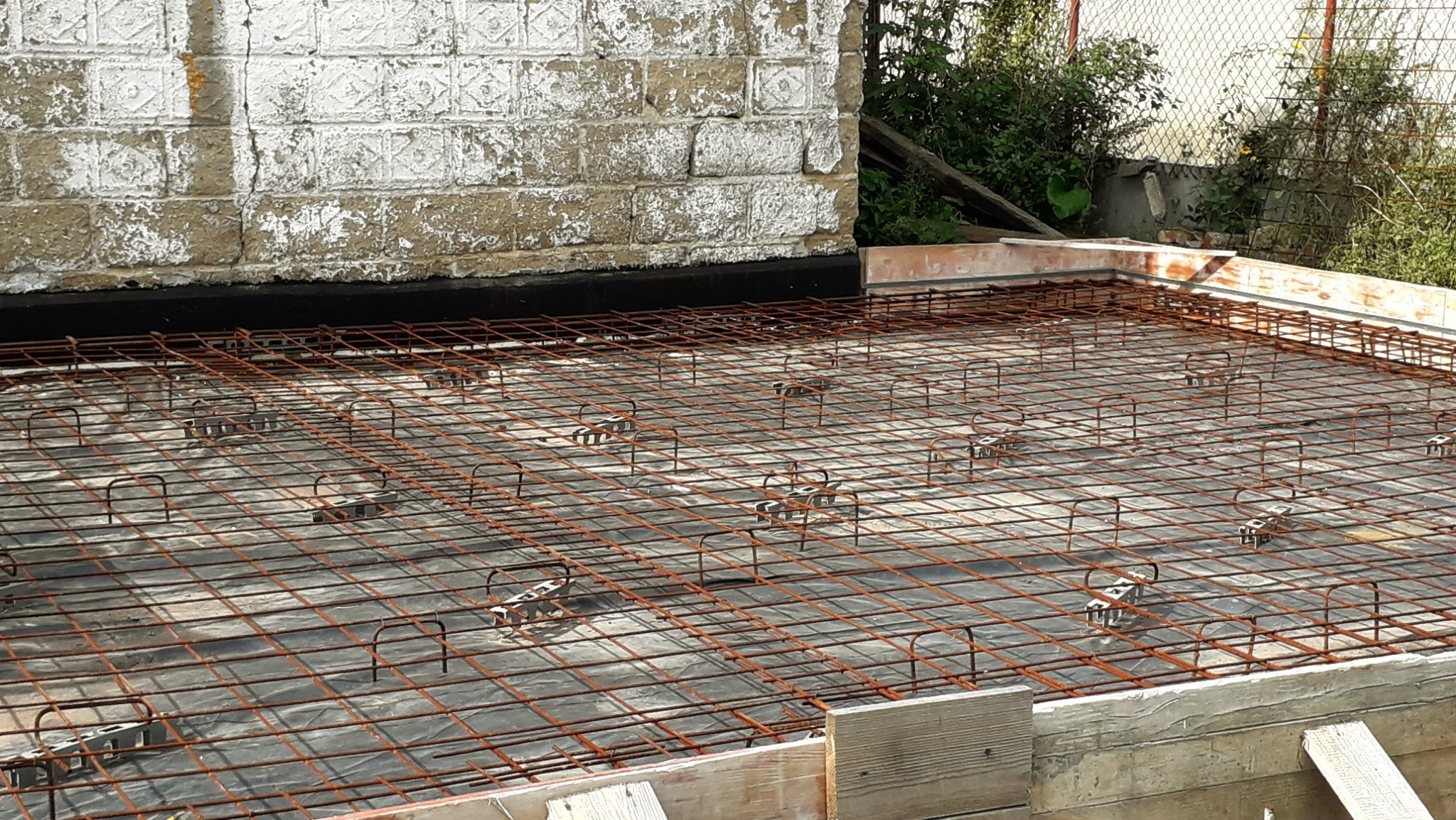 Zbrojenie płyty fundamentowej z użyciem dystansów, widoczne szalunki i izolacja. Ujęcie z góry, z widocznym murem w tle.