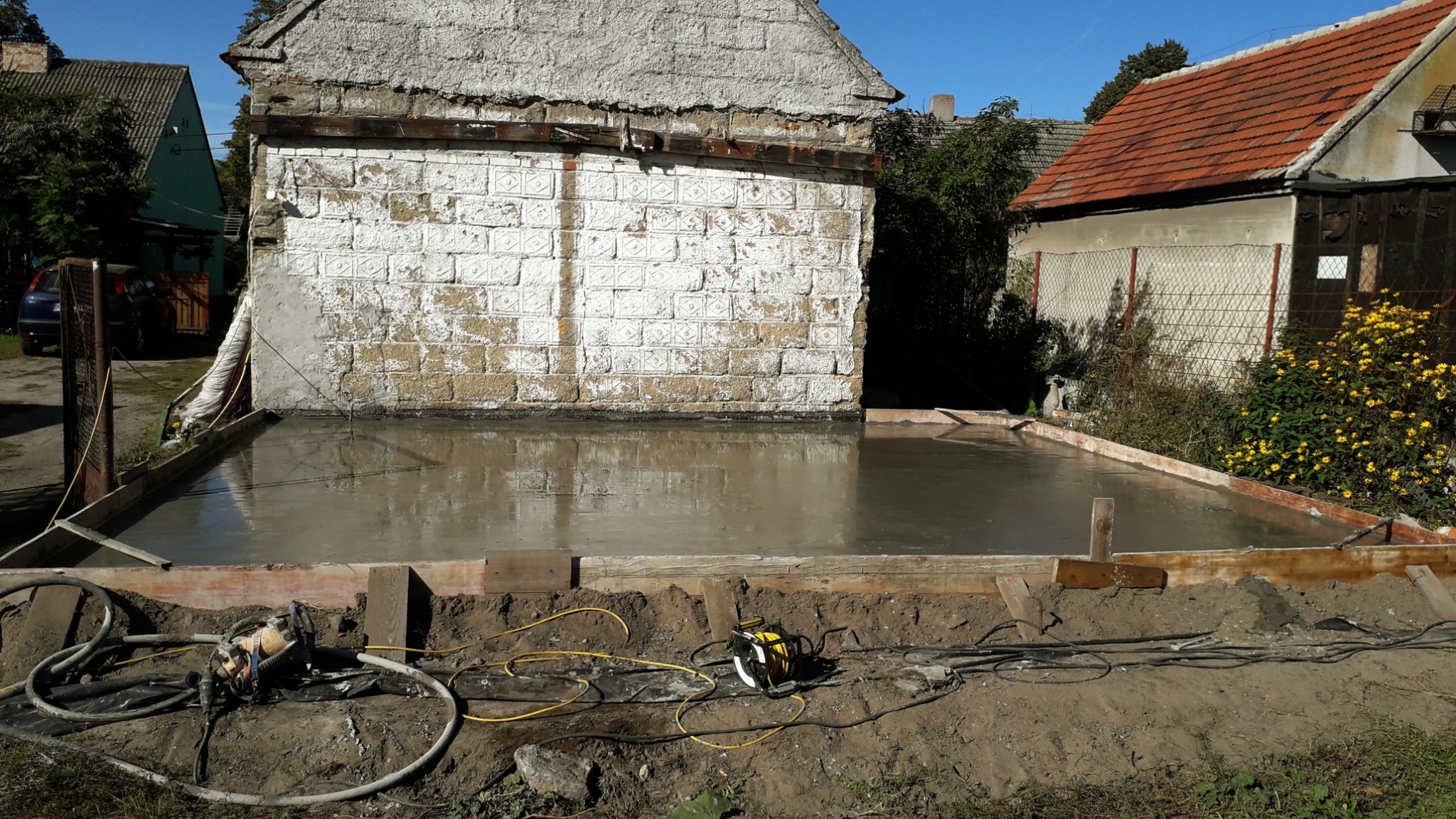 Wylany fundament pod budynek, z widocznym szalunkiem i narzędziami budowlanymi. Lustrzane odbicie nieba w mokrym betonie. W tle budynki mieszkalne.