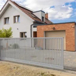 Nowoczesna, przesuwna brama wjazdowa z szarych paneli na tle domu z garażem obłożonym czerwoną cegłą i błękitnego nieba z chmurami.