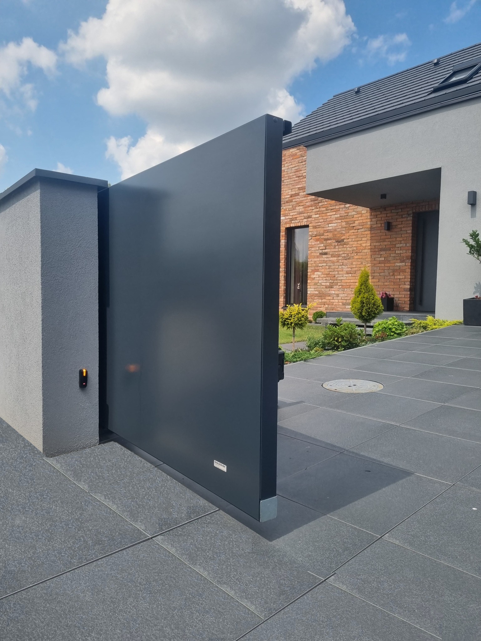 Nowoczesna, przesuwna brama wjazdowa w kolorze grafitowym, otwarta częściowo, z widoczną elektroniką sterującą na słupku, na tle domu z cegły i starannie zagospodarowanego ogrodu.