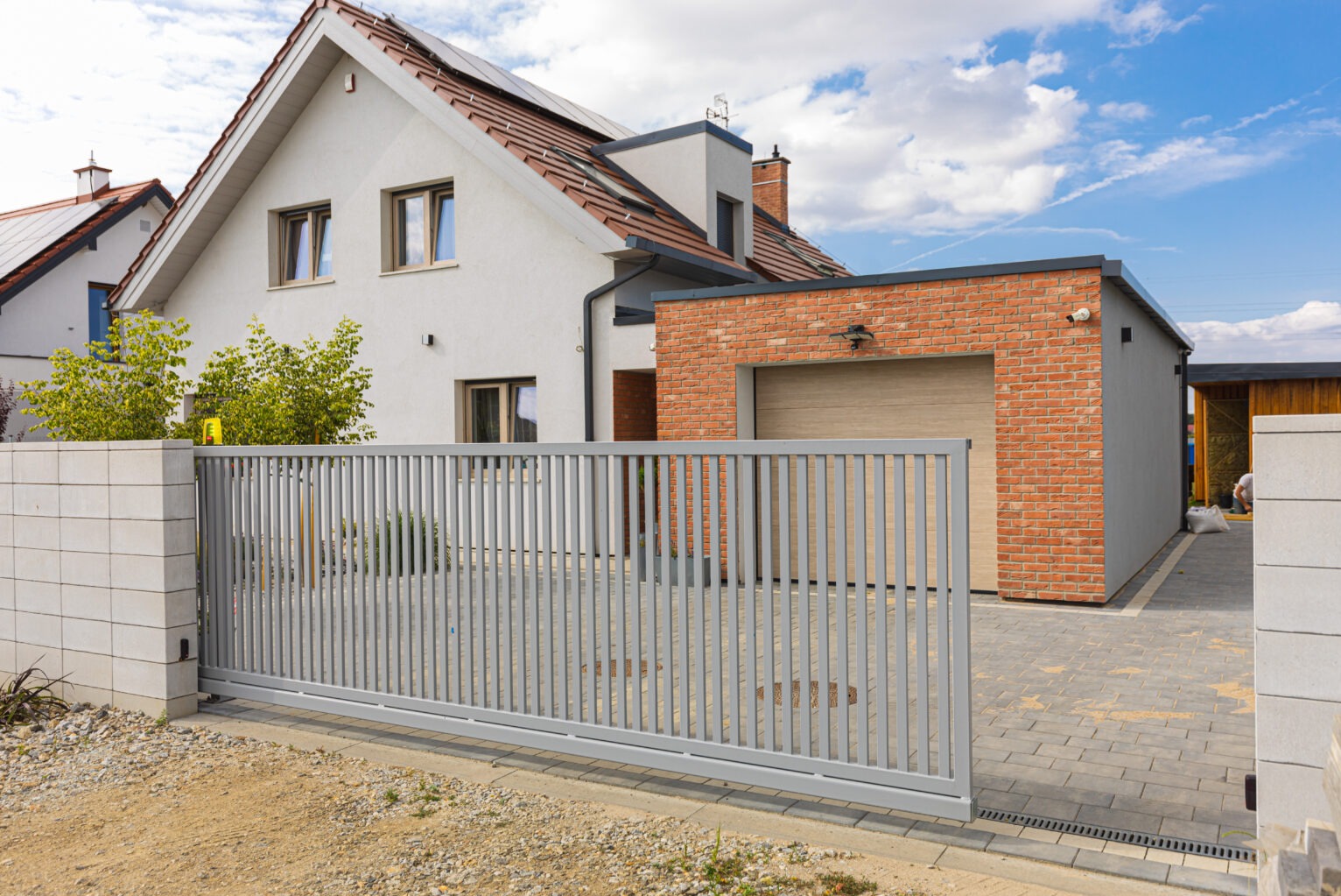Nowoczesna, przesuwna brama wjazdowa z szarych paneli na tle domu z garażem obłożonym czerwoną cegłą i błękitnego nieba z chmurami.