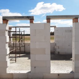 GZB - Kompleksowe prace ziemne, murarskie - Wznoszone ściany domu z betonu komórkowego Ytong, widoczne nadproża z napisem 'POZBRUK' i metalowe rusztowanie wewnątrz, w tle łąka pod błękitnym niebem.