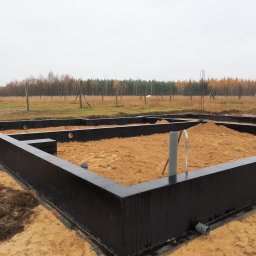 GZB - Kompleksowe prace ziemne, murarskie - Fundamenty z bloczków pomalowane na czarno, zasypane piaskiem, widoczne rury kanalizacyjne i wystające pręty zbrojeniowe na tle jesiennego krajobrazu.